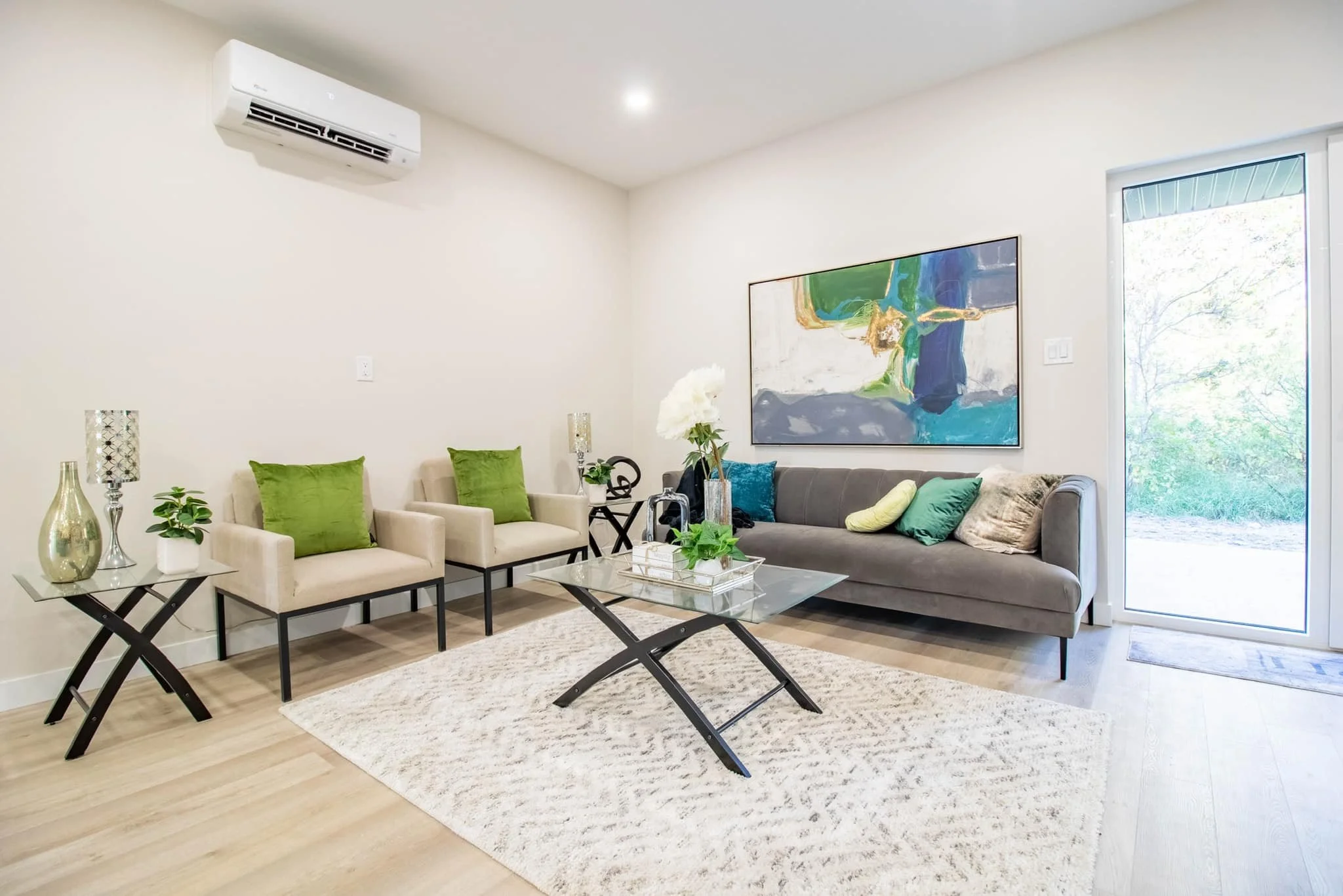 Living room with beige chairs, gray sofa, glass coffee table, and artwork on the wall; natural light from a sliding door.