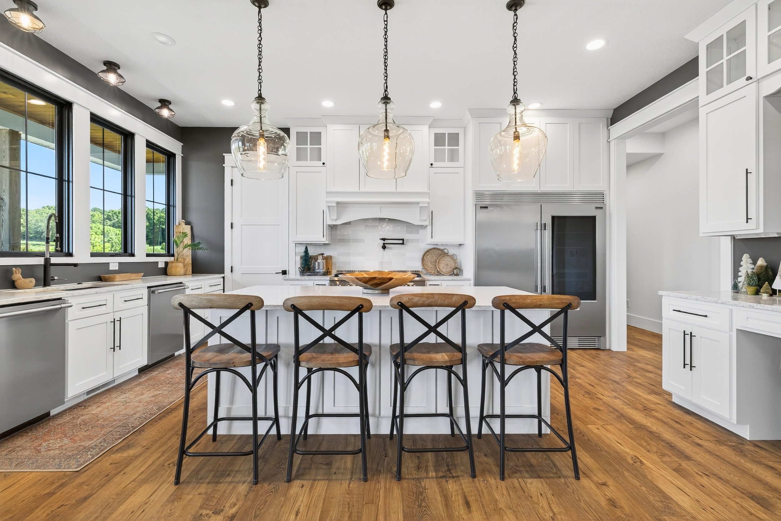 Modern kitchen with white cabinets, a large island with four bar stools, stainless steel appliances, and pendant lighting, hardwood floors, and large windows showing outdoor greenery.
