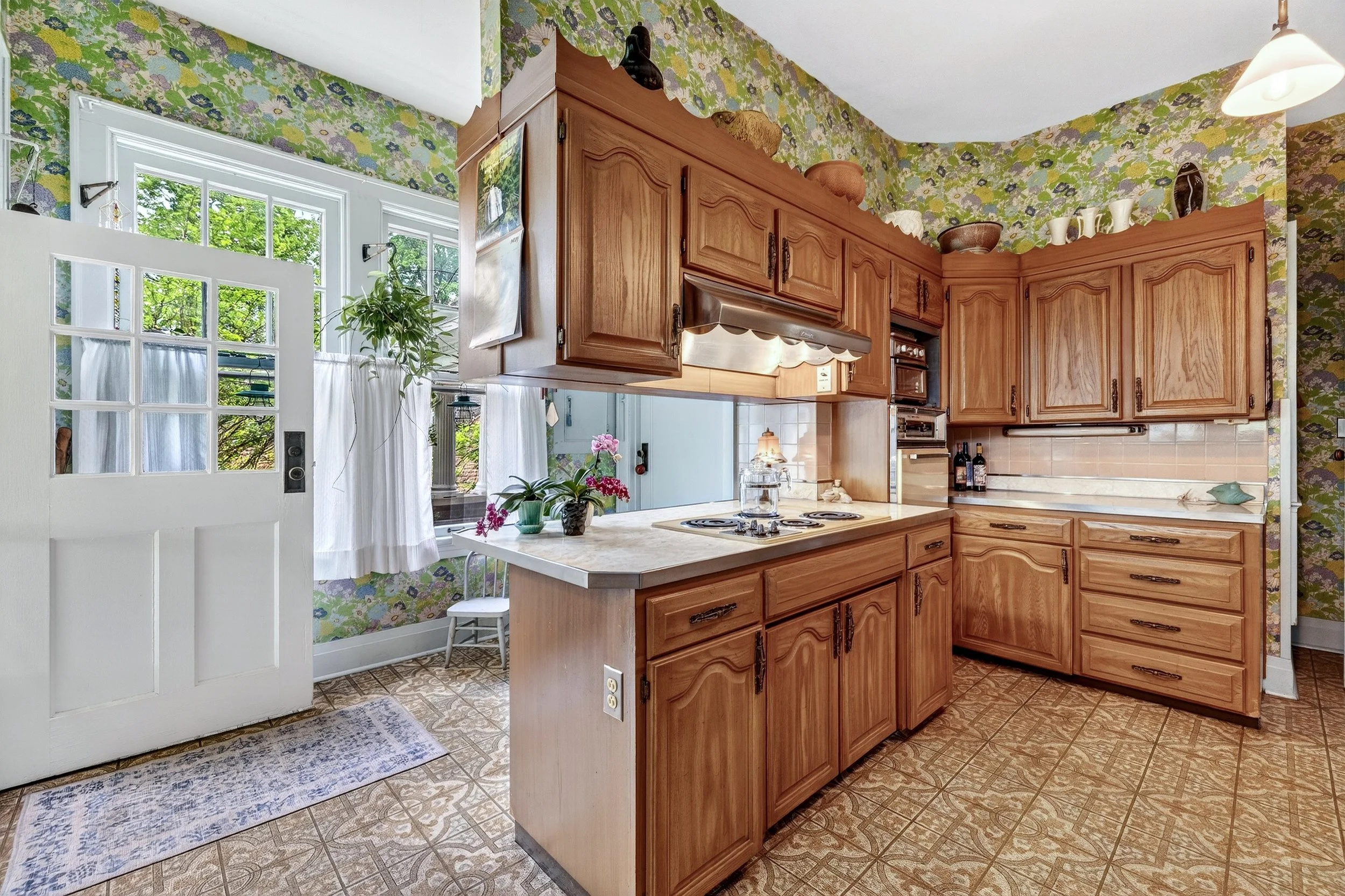 A vintage-style kitchen with wooden cabinets, floral wallpaper, and a tiled linoleum floor. There are windows with white curtains, a potted plant, a small table, and some decorative items on top of the cabinets.