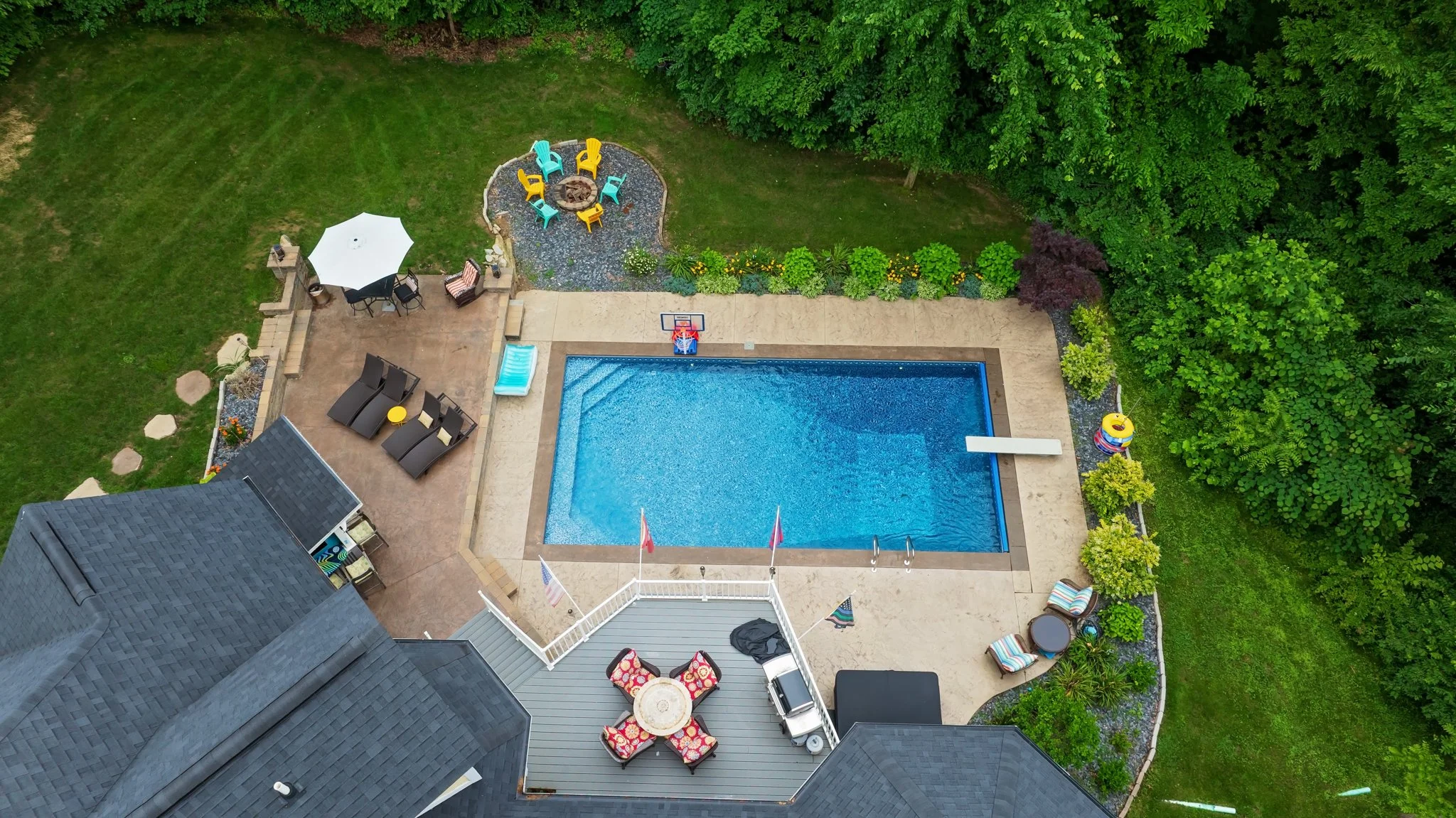 A backyard with a swimming pool, outdoor seating area, children's play area with a slide and ring toss game, surrounded by green trees and shrubs.