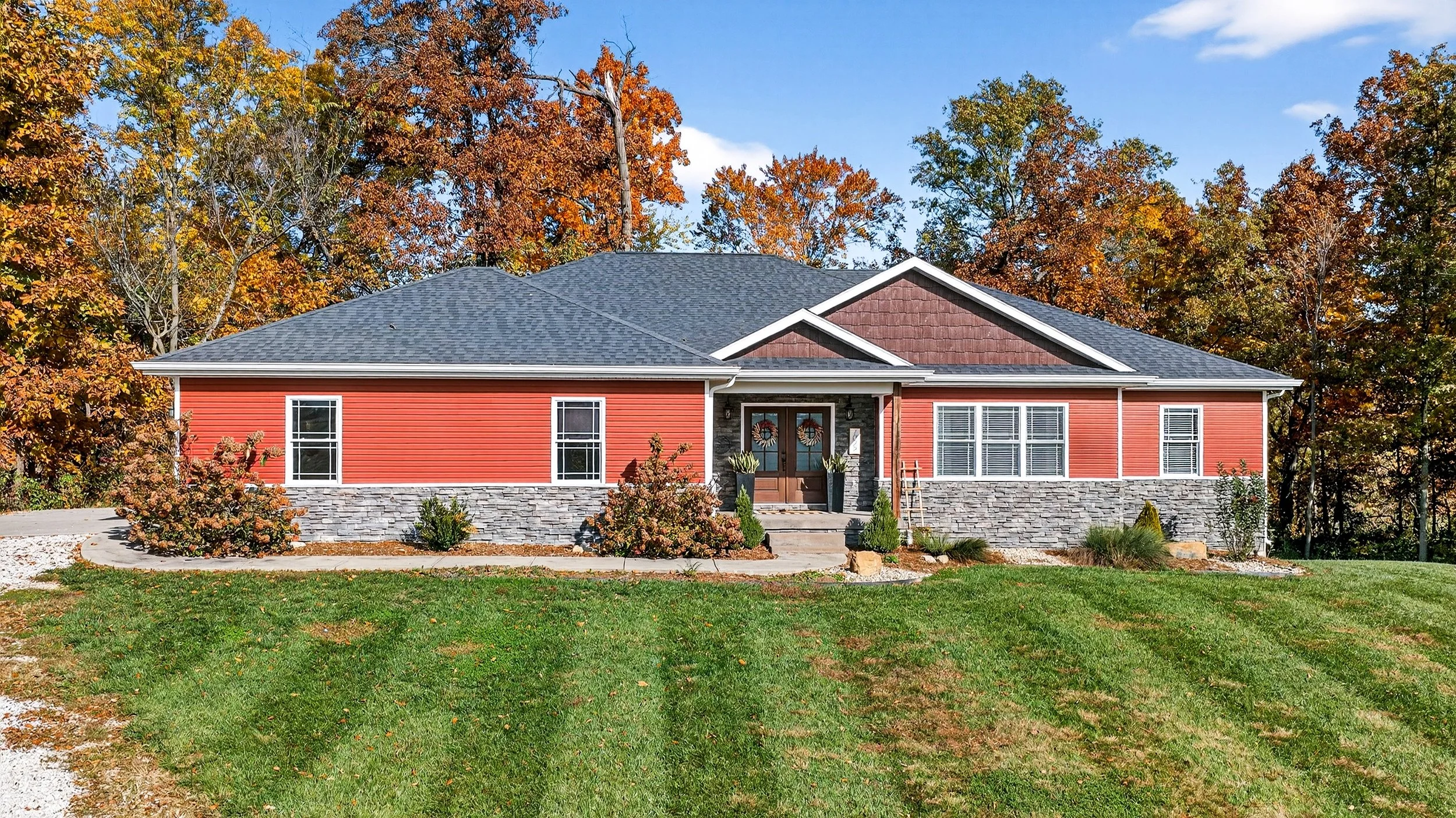 Single-story house with red siding, stone foundation, and a dark gray roof, surrounded by trees with autumn foliage, a front yard with grass, bushes, and a concrete walkway leading to the front door.