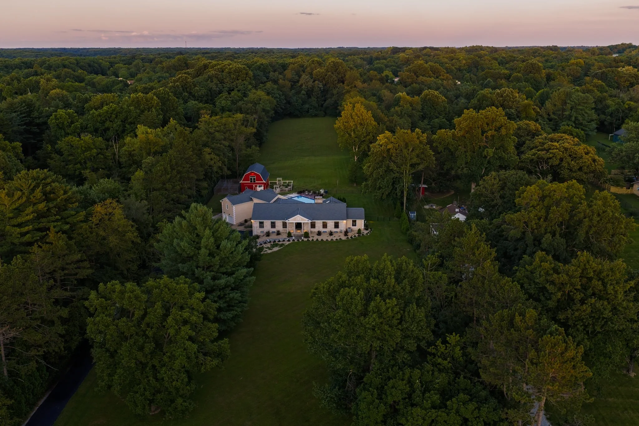 A large house with a covered porch and a backyard swimming pool, surrounded by dense trees, on a spacious property at sunset