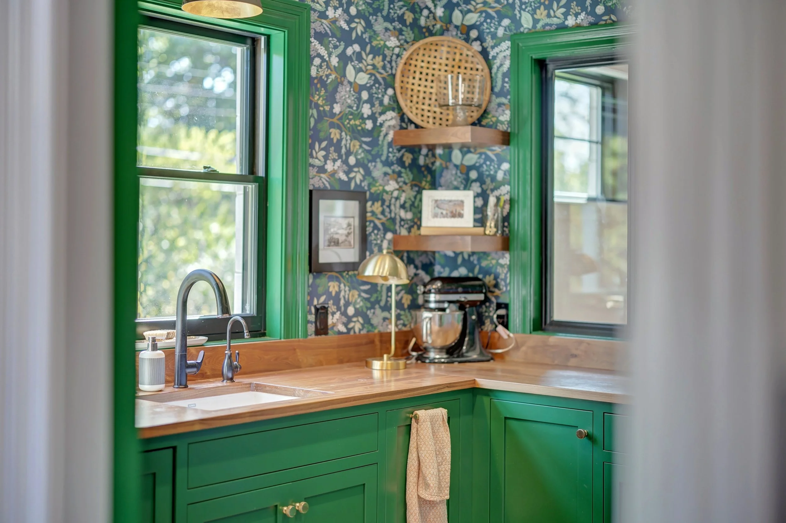 Kitchen with green cabinets and window frames, floral wallpaper, wooden countertop, brass desk lamp, stand mixer, and wall shelves with decorative items.