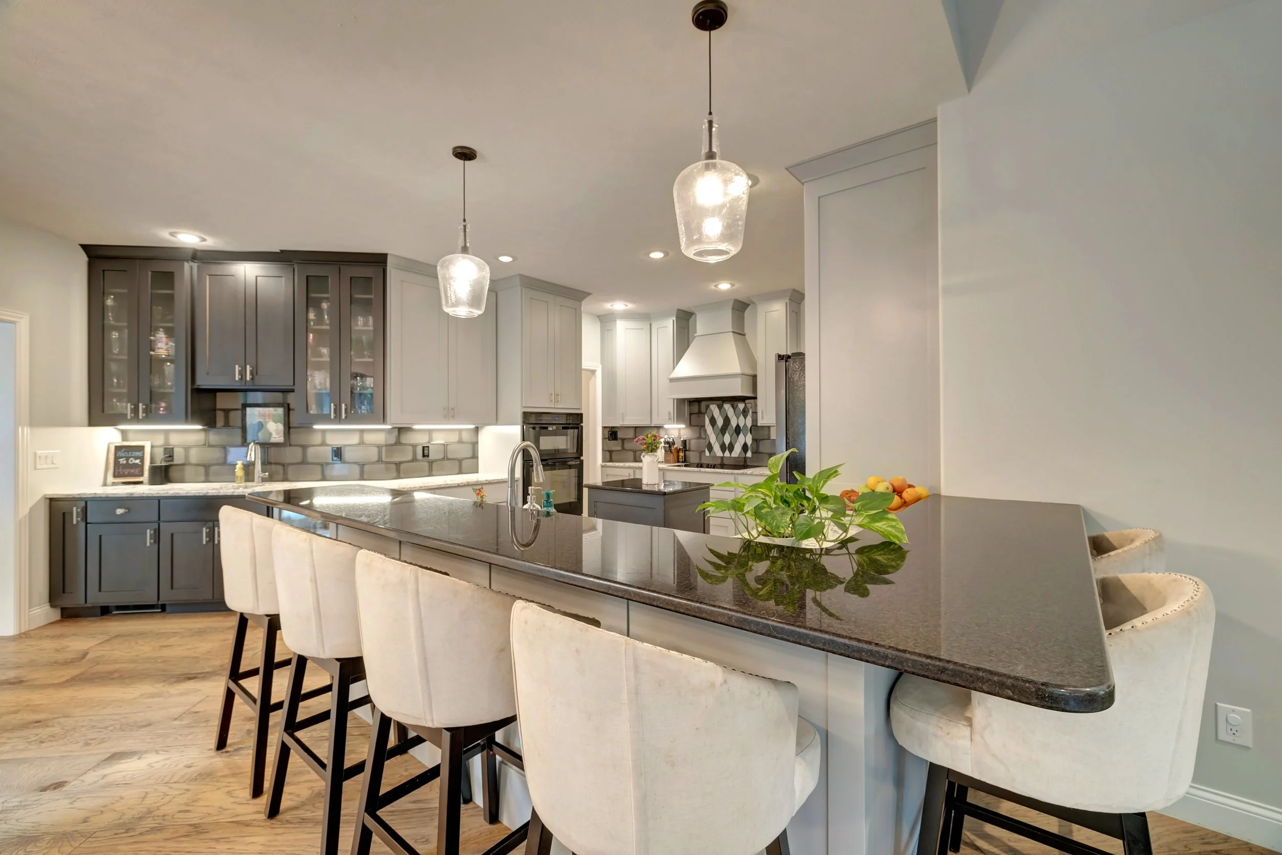 Modern kitchen with gray and white cabinetry, black granite countertops, a large island with beige chairs, hanging pendant lights, and hardwood floors.