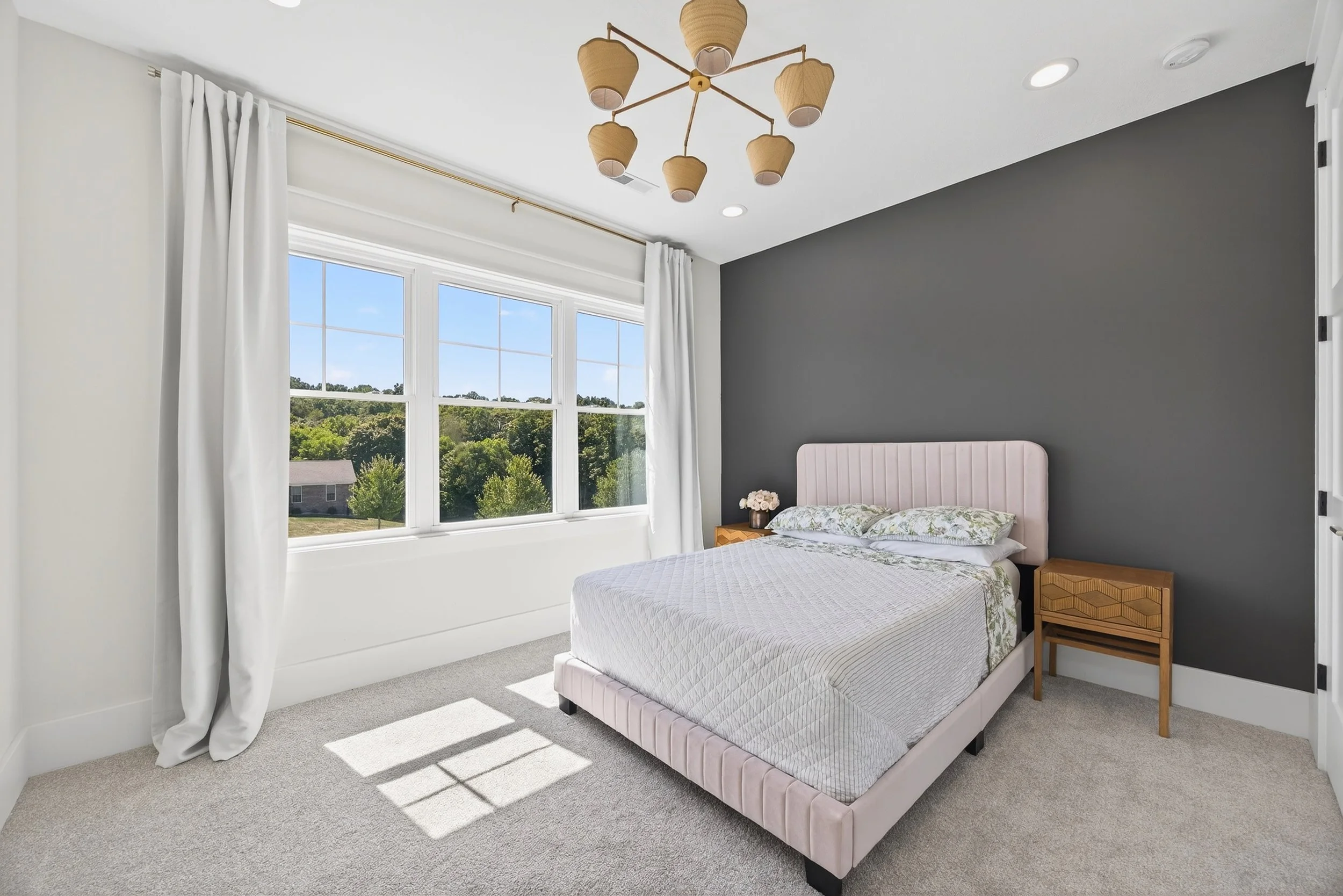 Bedroom with large window, white curtains, pink upholstered bed, gray accent wall, wooden nightstands, and a ceiling light fixture.