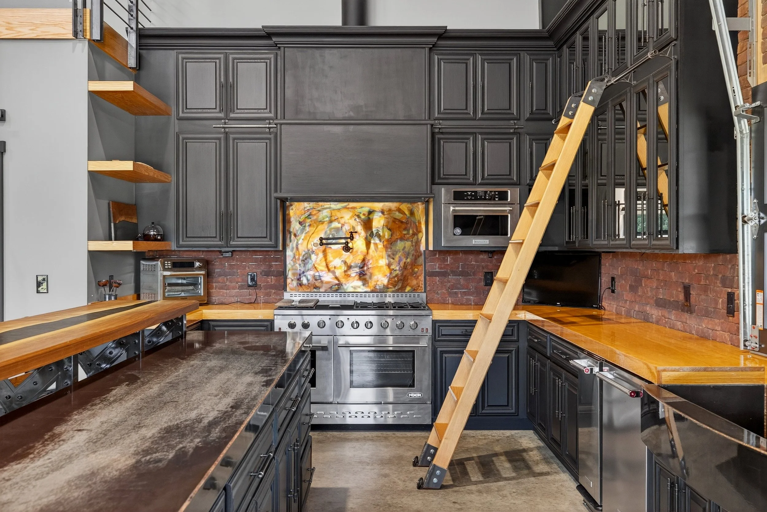 Modern kitchen with dark gray cabinets, brick backsplash, wooden countertops, a ladder, and stainless steel appliances.