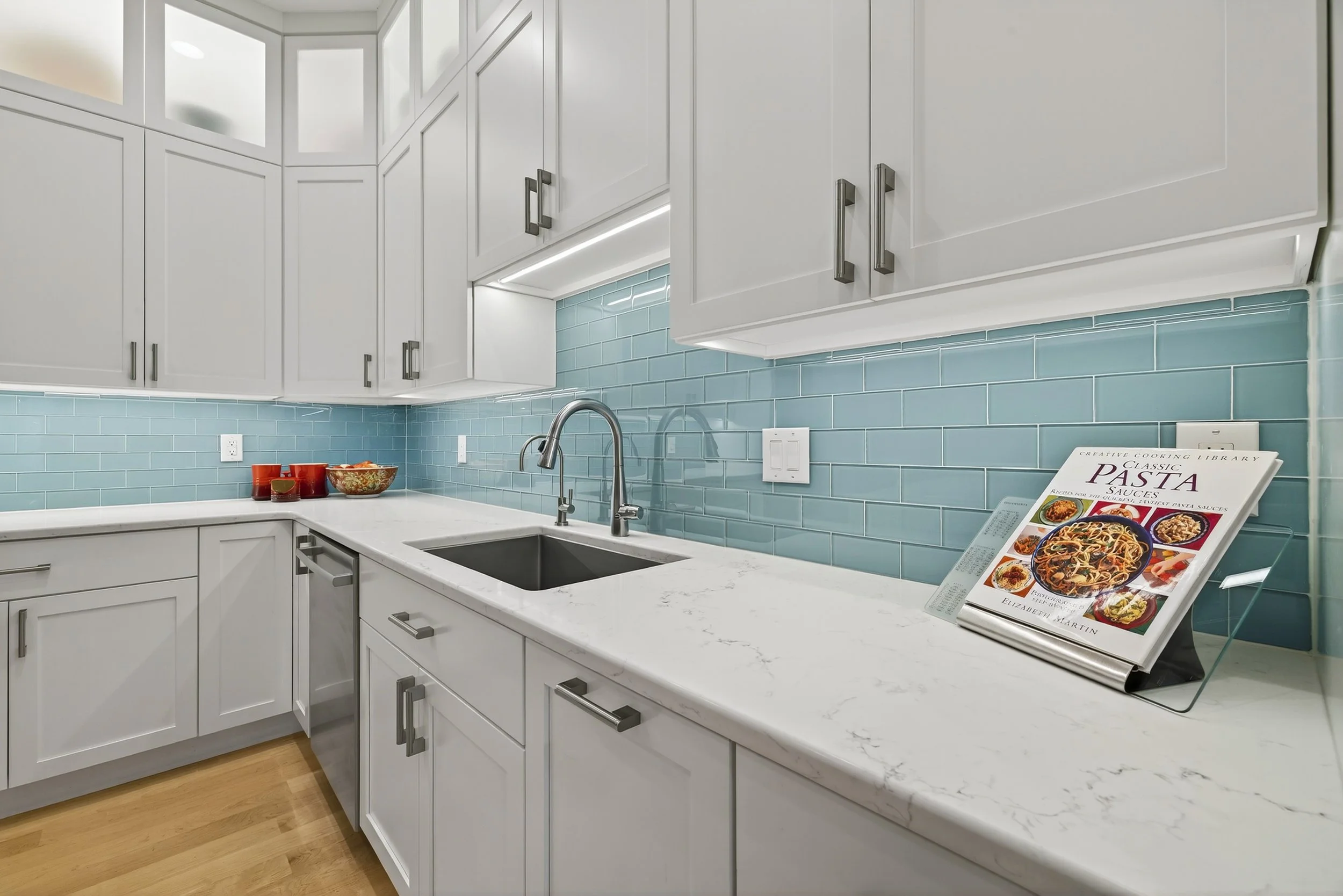 Modern kitchen with white cabinets, marble countertop, blue subway tile backsplash, and a cookbook stand holding a pasta cookbook.