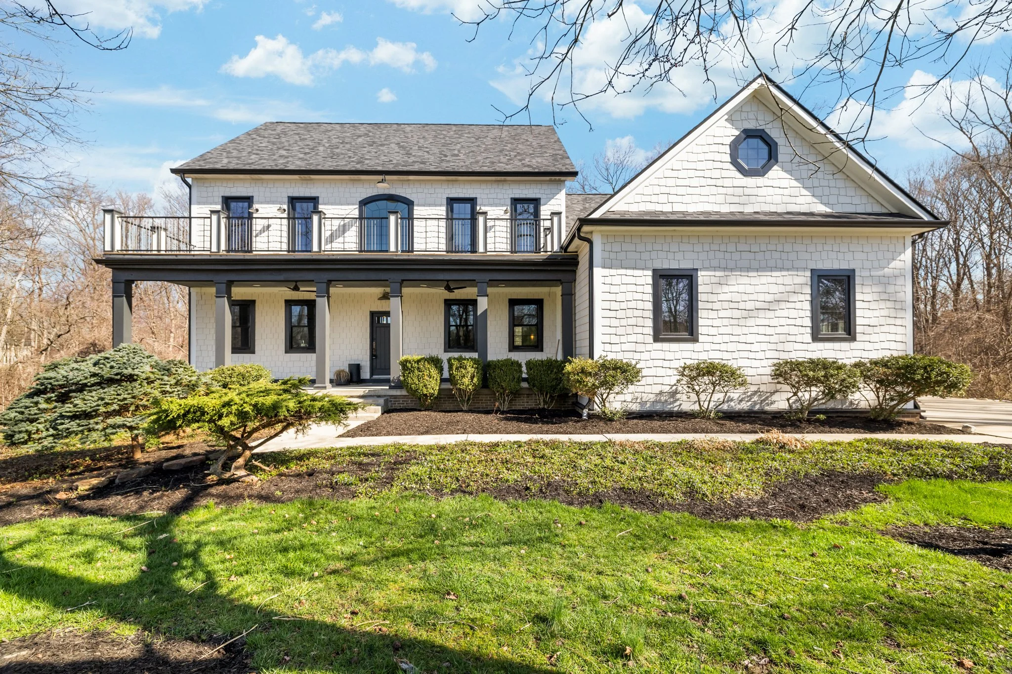 Front view of a two-story white house with black window frames and a balcony, surrounded by a landscaped yard with bushes and grass, under a partly cloudy sky.