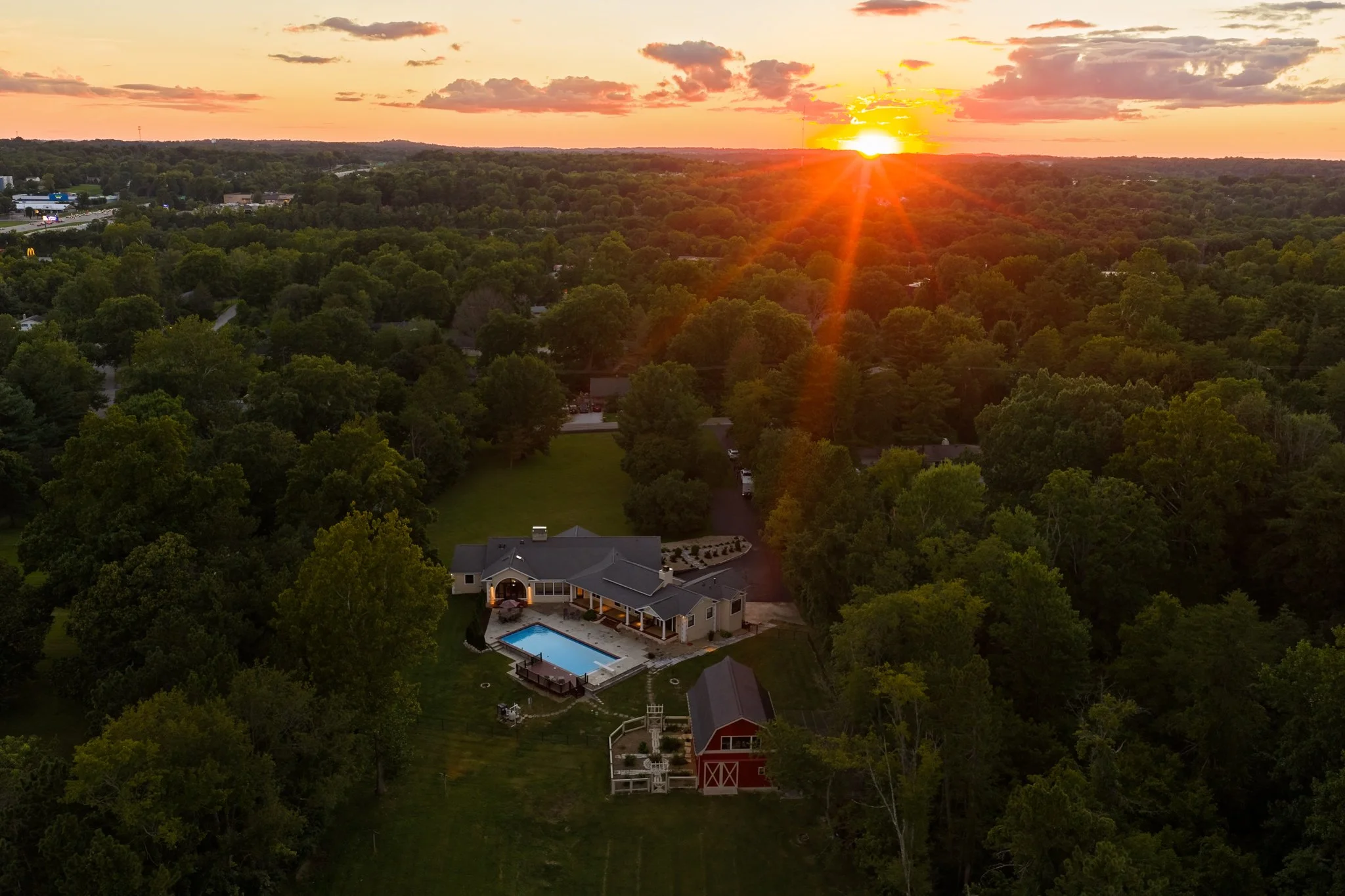 Aerial view of a suburban neighborhood at sunset, showing a house with a swimming pool, surrounded by trees and open land.