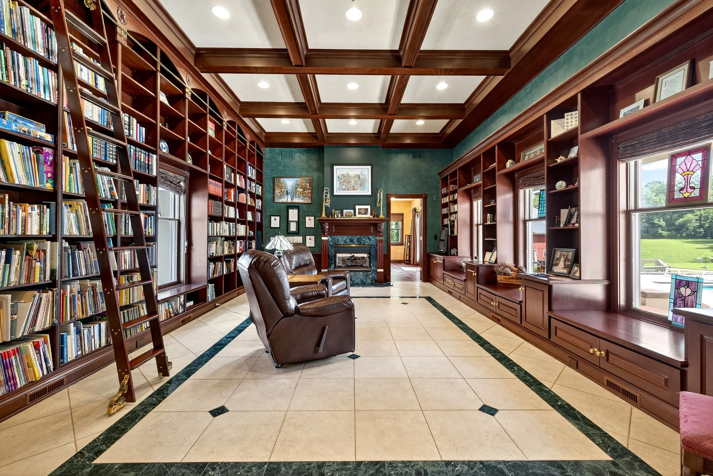 A spacious home library with dark wood built-in bookshelves on all sides, filled with books. There are large windows with stained glass accents and seating including a leather recliner. The room features a tiled floor and a decorative coffered ceilin