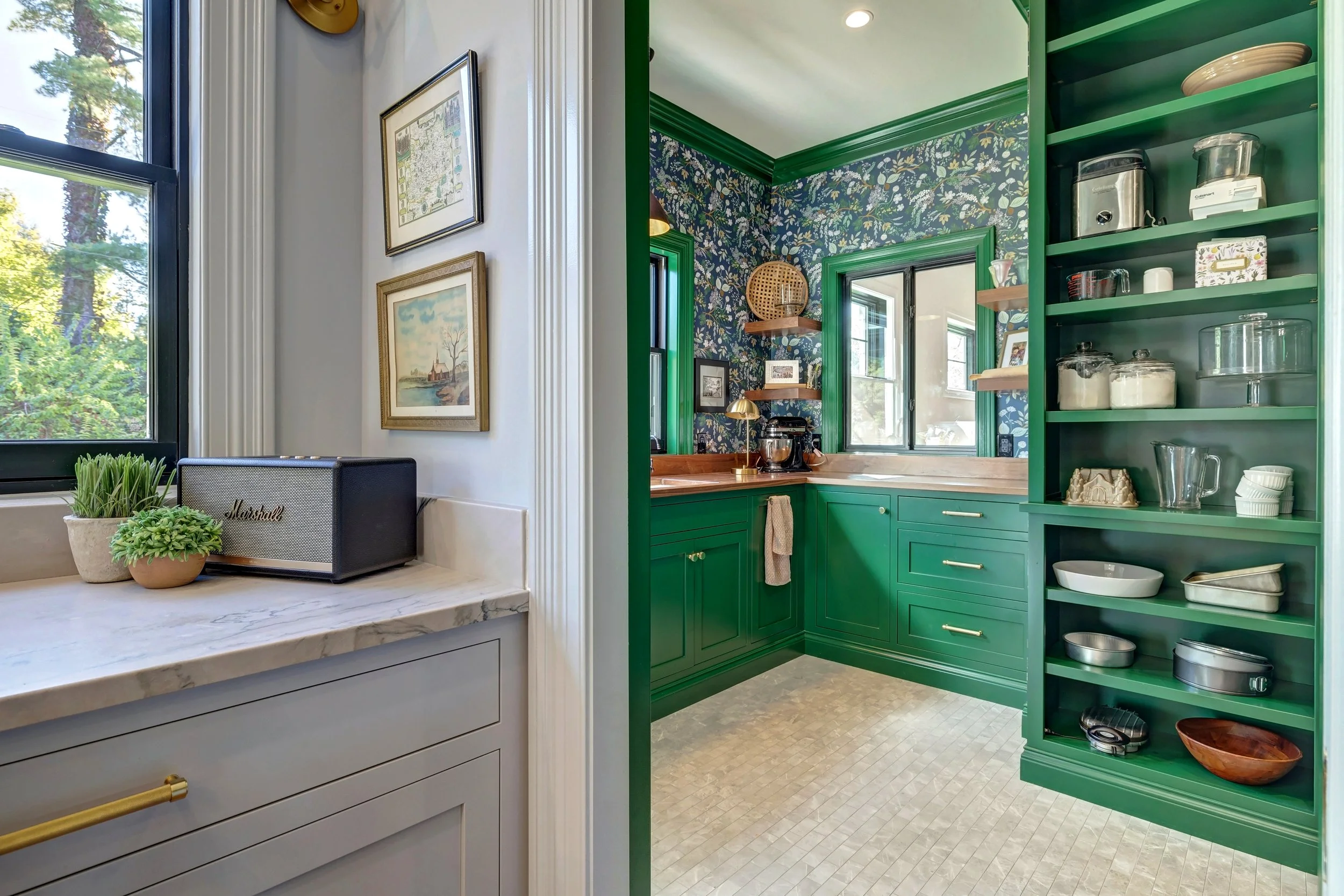 A view into a kitchen with green cabinetry, floral wallpaper, and open shelves with kitchenware. The adjacent room has a marble countertop, window with plants, and framed artwork on the wall.