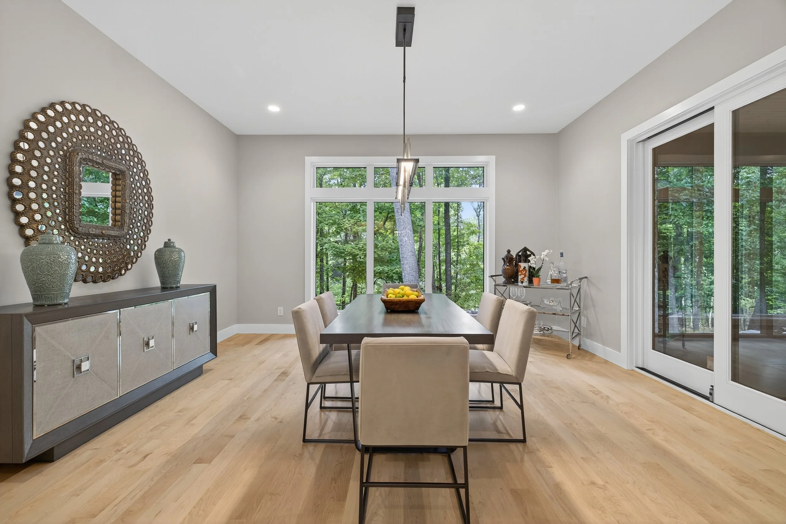 A bright dining room with a wooden table, six beige chairs, a sideboard with decorative vases, and large windows overlooking a green wooded area.