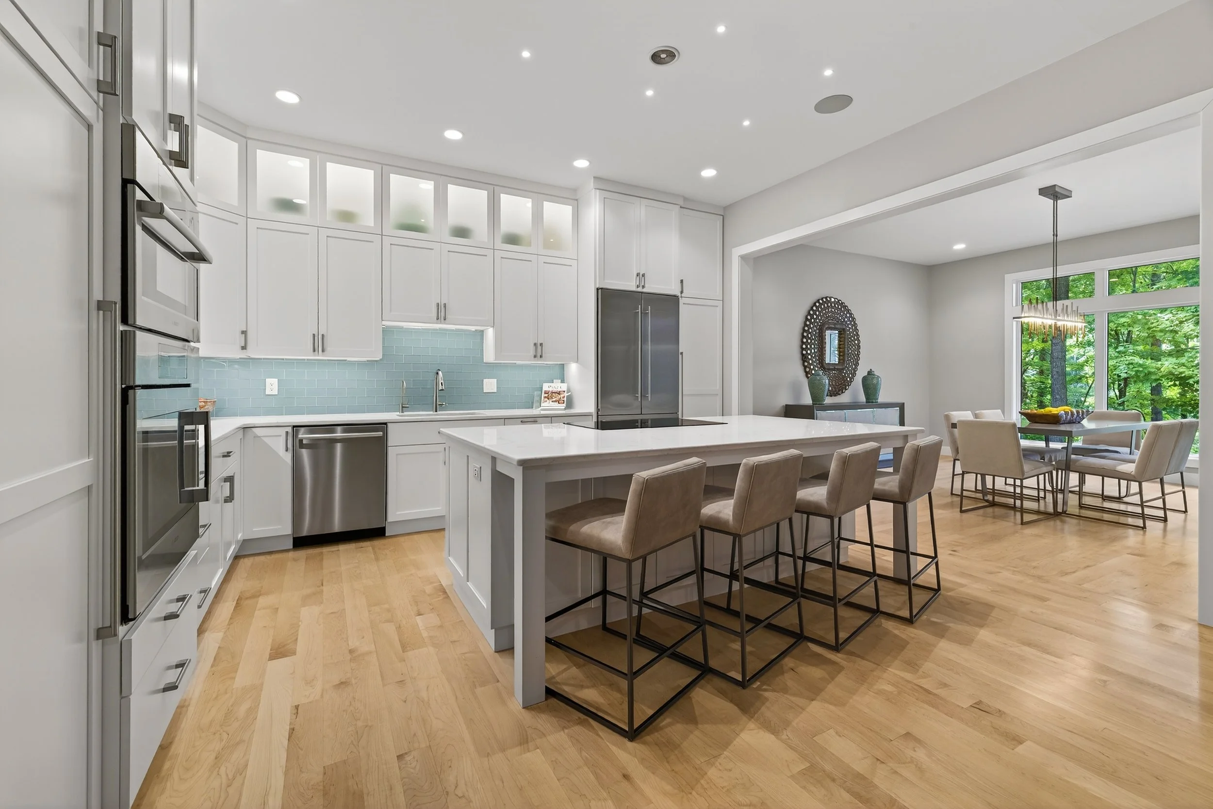 Modern kitchen with white cabinets, light blue subway tile backsplash, stainless steel appliances, a large kitchen island with beige bar stools, and a dining area with a round table and six beige chairs next to large windows with green trees outside.