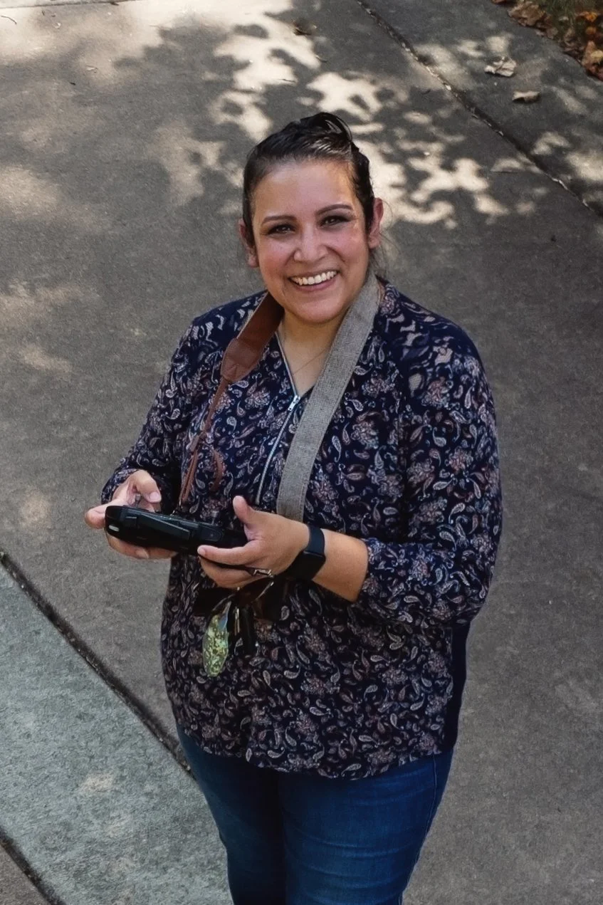 A smiling woman with dark hair in a ponytail, wearing a patterned blouse, jeans, and a watch. She is holding a smartphone and standing on a sidewalk with shadows of trees overhead.
