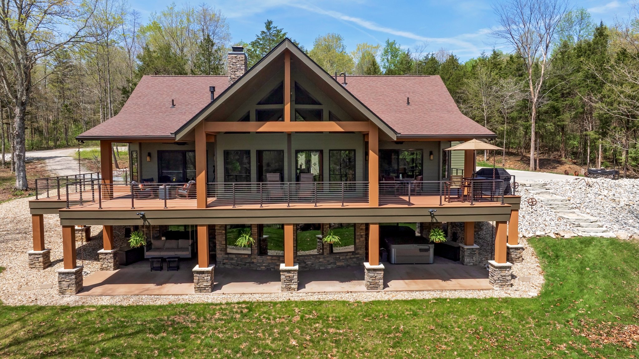 A large two-story house with a spacious wrap-around deck, surrounded by trees and a green lawn, with outdoor furniture and a grill on the deck.