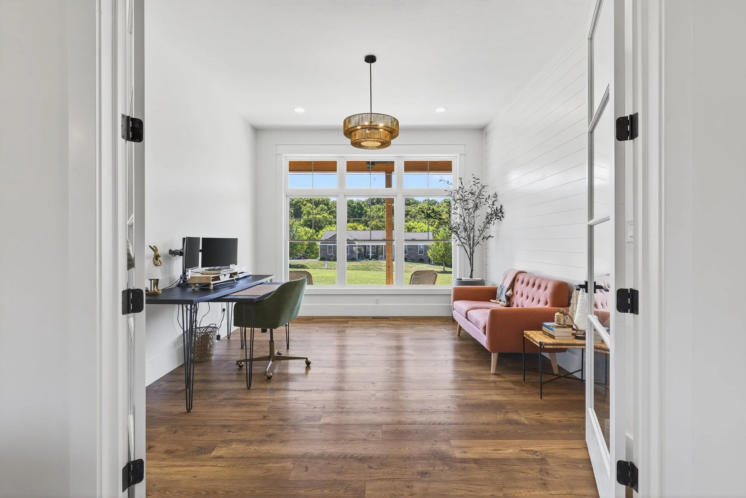 Bright living room with large window, pink sofa, green desk chair, small table, and wooden flooring.