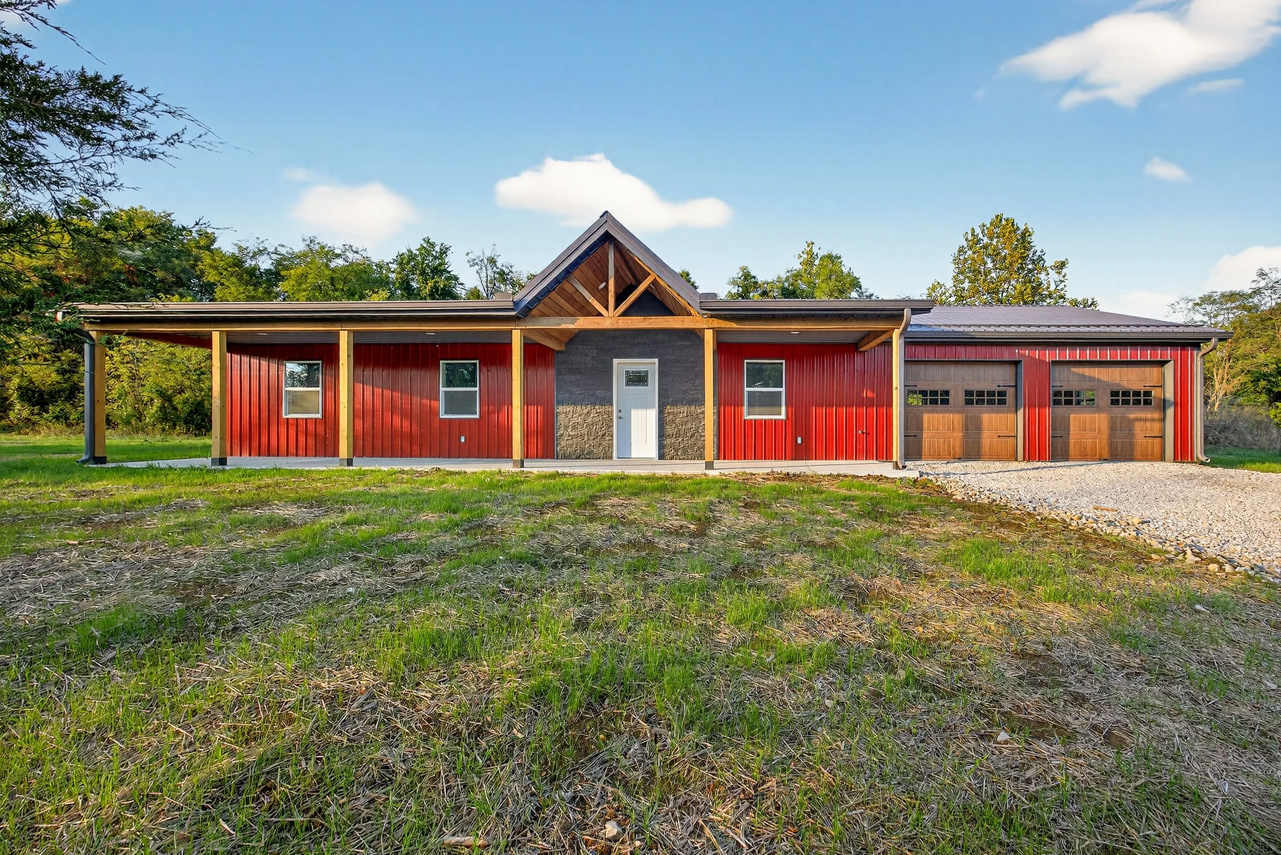Front view of a modern farmhouse with red siding, a gable roof, and a two-car garage, surrounded by a grassy yard and trees under a blue sky.