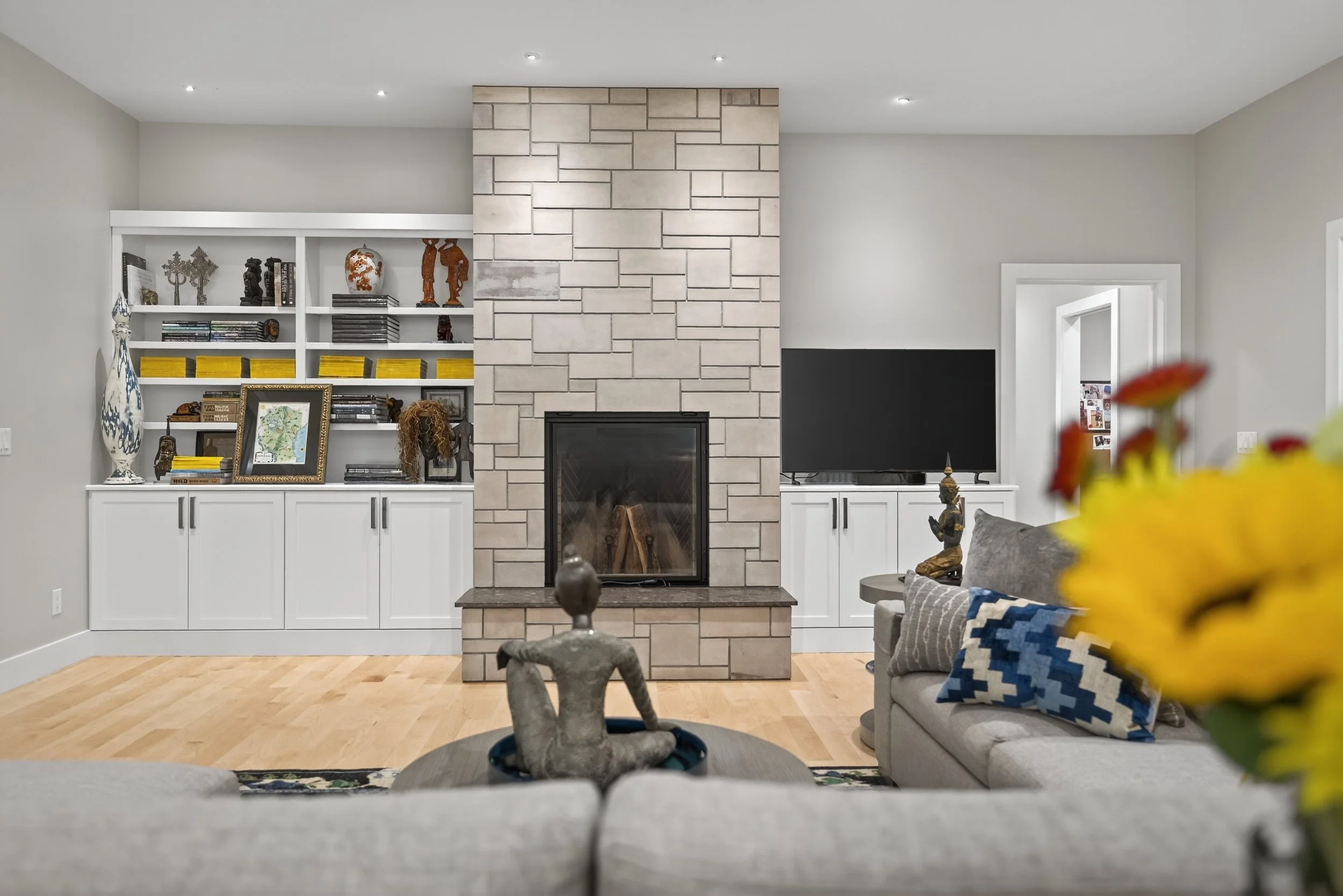 Living room with a fireplace, built-in white shelves filled with books and decorative items, a flat-screen TV, and a beige sofa with patterned pillows, a small gray side table, and a yellow flower in the foreground.