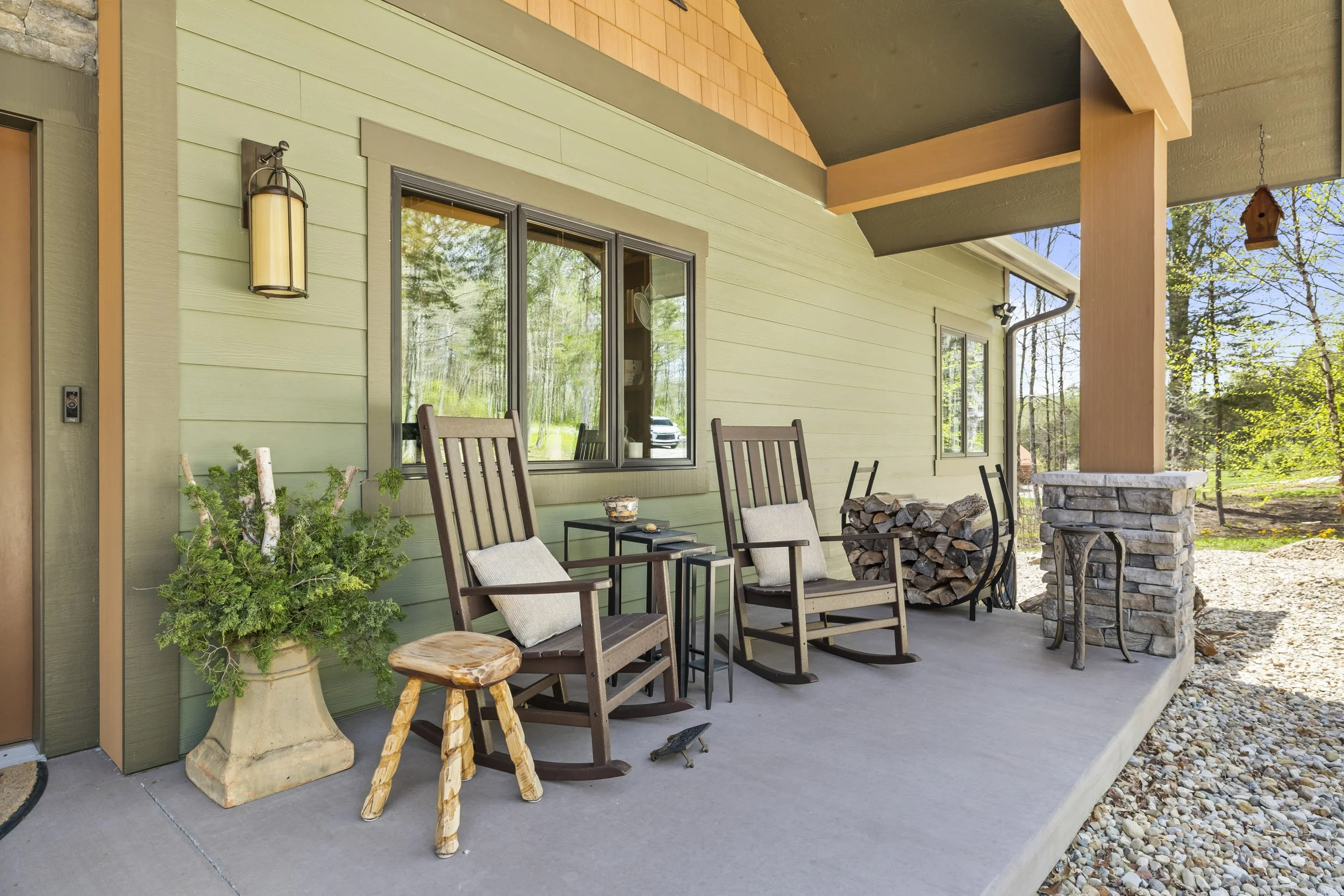 Outdoor porch with two wooden rocking chairs and a small side table, decorated with pillows, in front of a green house with large windows and surrounded by trees.