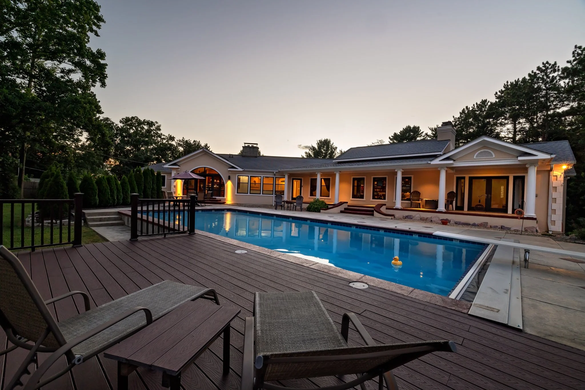 Backyard with a swimming pool, poolside furniture, and a large house with a covered porch, illuminated at dusk.