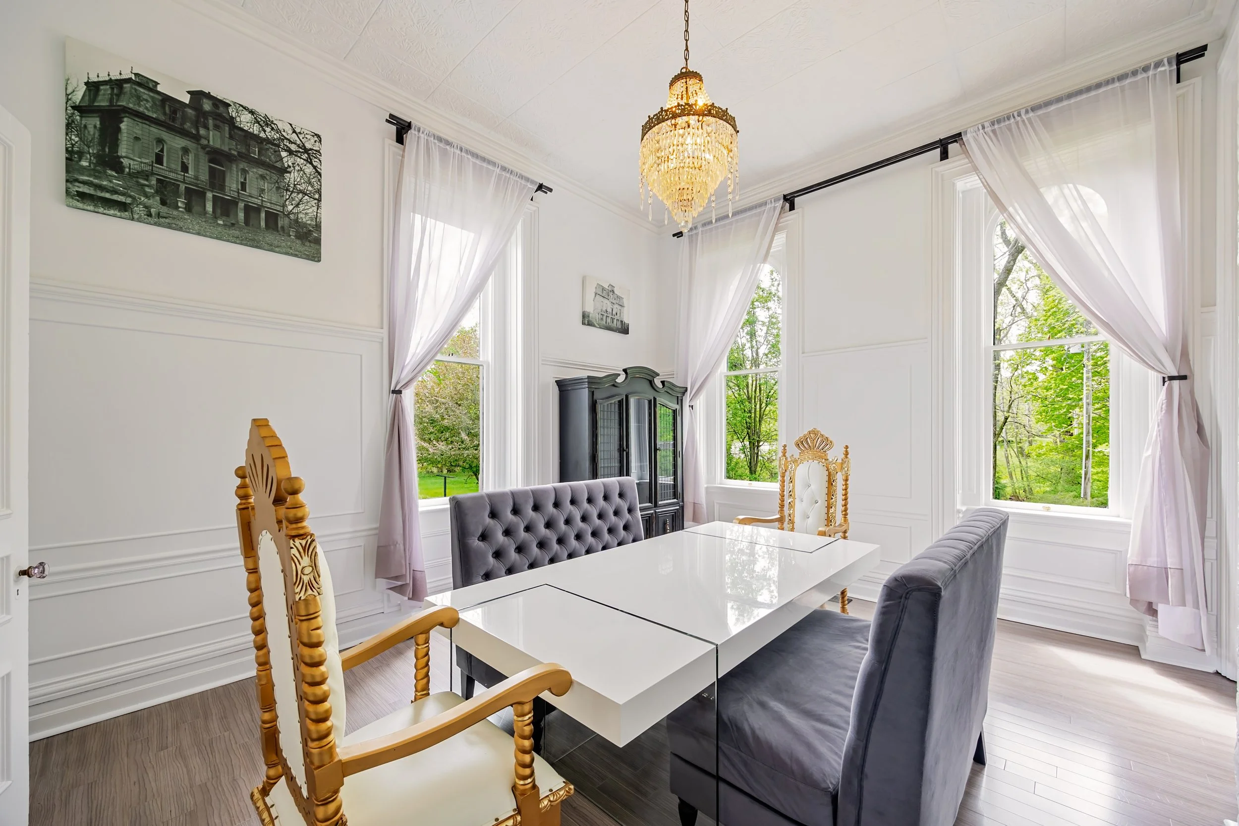Bright dining room with white walls, large windows with sheer curtains, a crystal chandelier, a black china cabinet, and a white table with various chairs, including a black tufted and two wooden armchairs.
