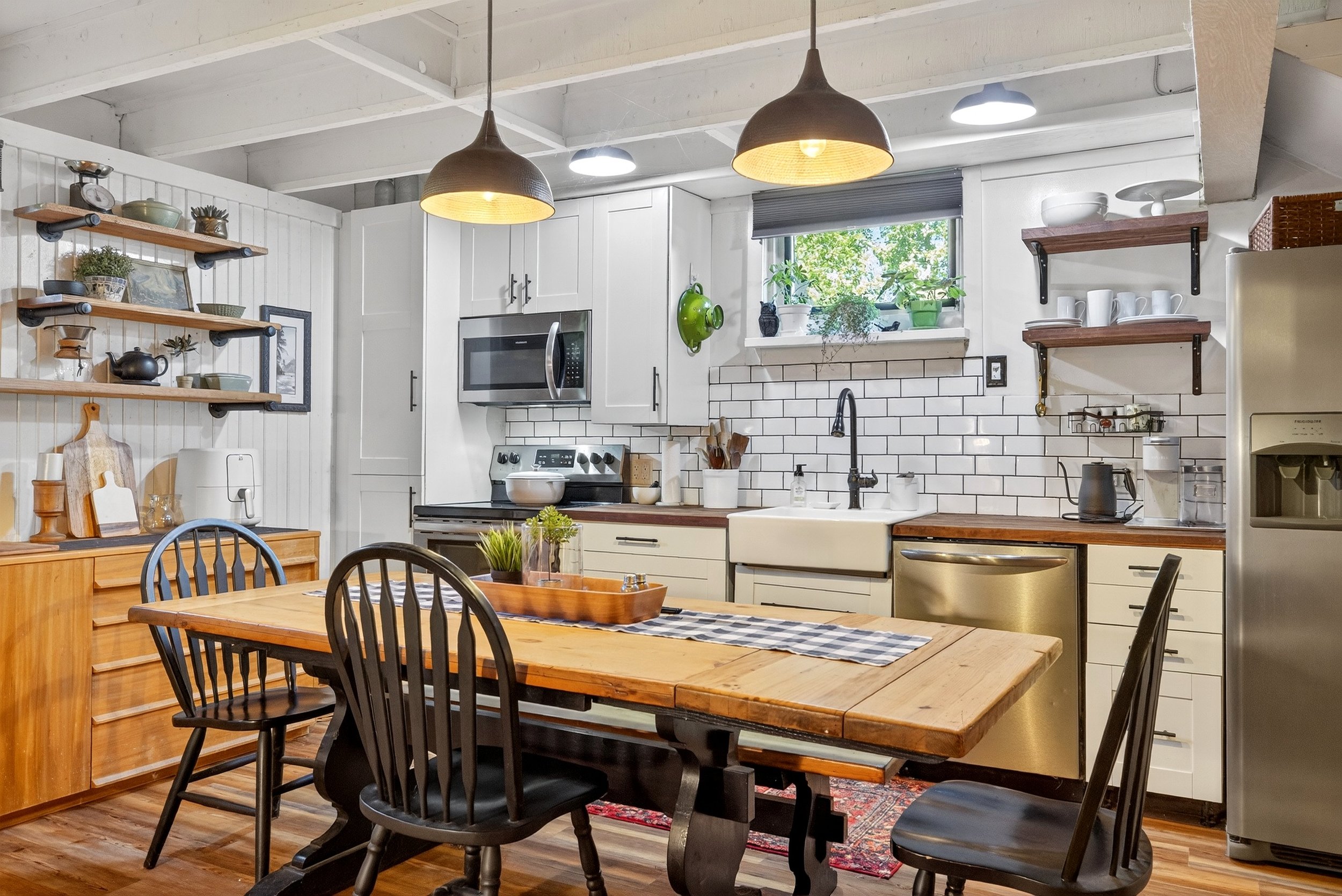 Rustic kitchen with white cabinetry, black appliances, open wooden shelves, a wooden dining table with black chairs, pendant lighting, and potted plants on a window sill.