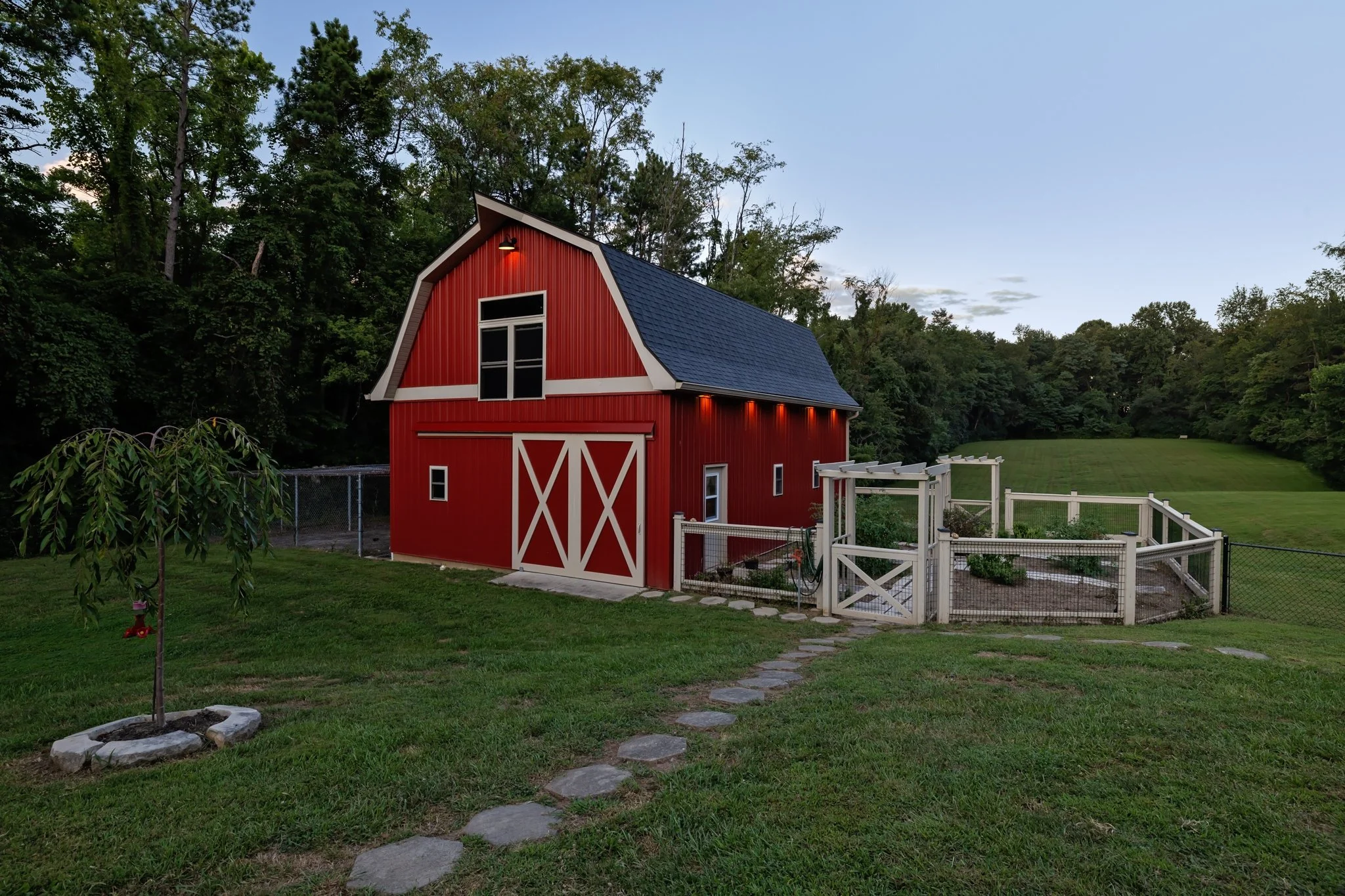 A red barn with white trim and a blue roof in a grassy backyard with trees in the background, at dusk.