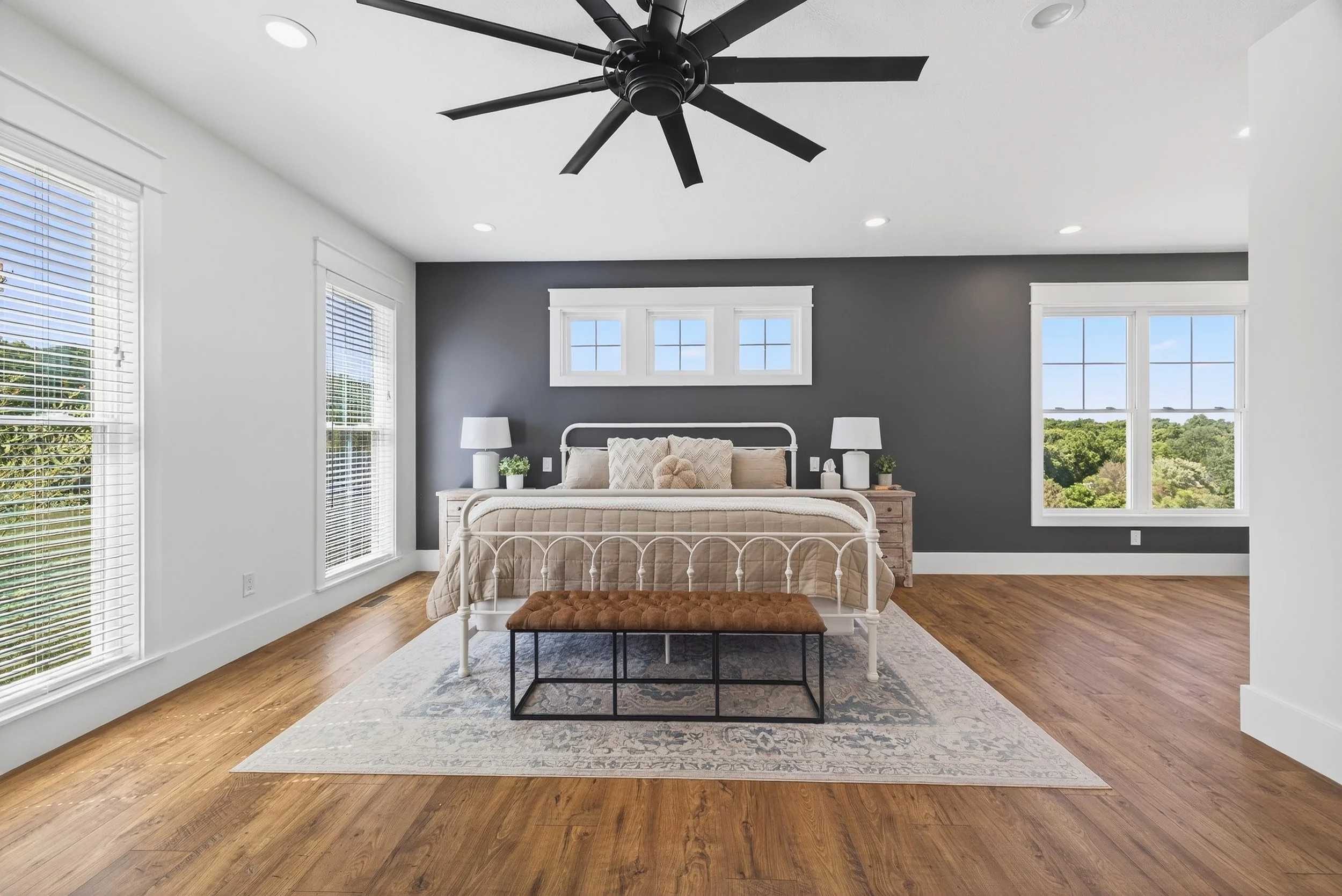 Bright bedroom with dark accent wall behind bed, white bed frame, beige bedding, and wooden nightstands with lamps. Large windows on two sides with views of greenery, ceiling fan overhead, hardwood floors, and a patterned area rug under the bed.
