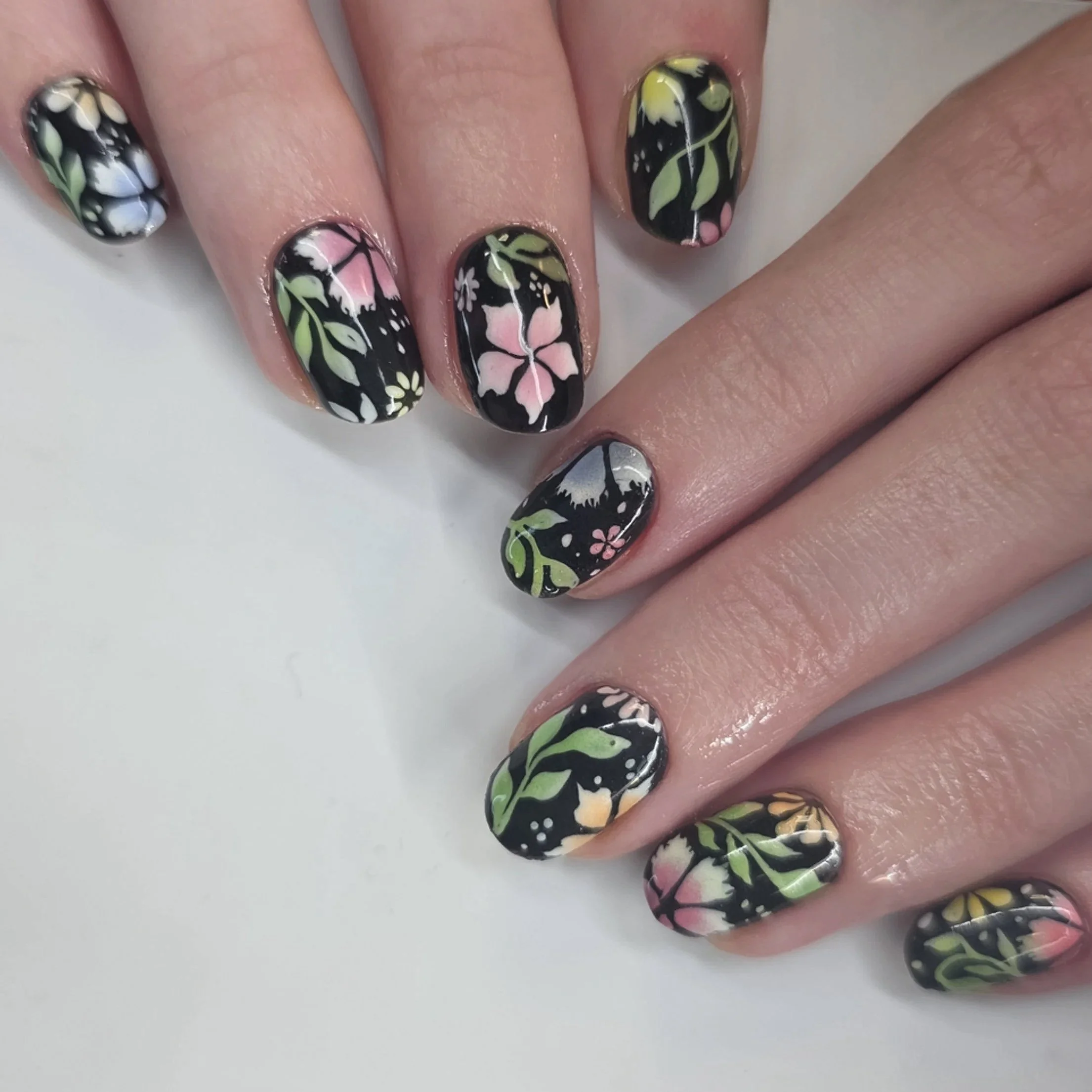 Colorful floral nail art on black painted nails with pink, yellow, green, and white flower and leaf designs.