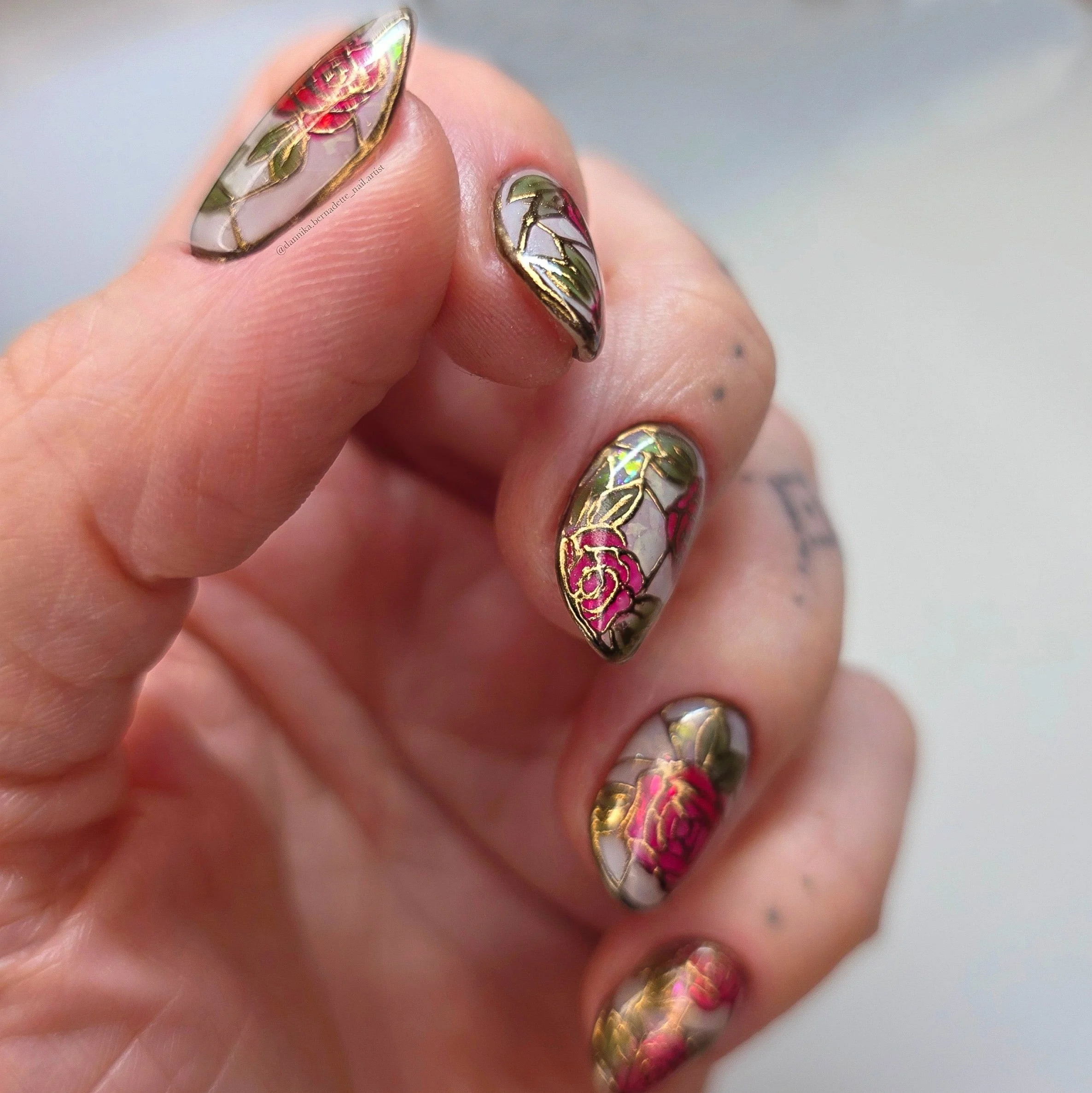 Close-up of a hand with nails featuring stained glass floral nail art with pink, red, white, and green colors, and gold outlines.