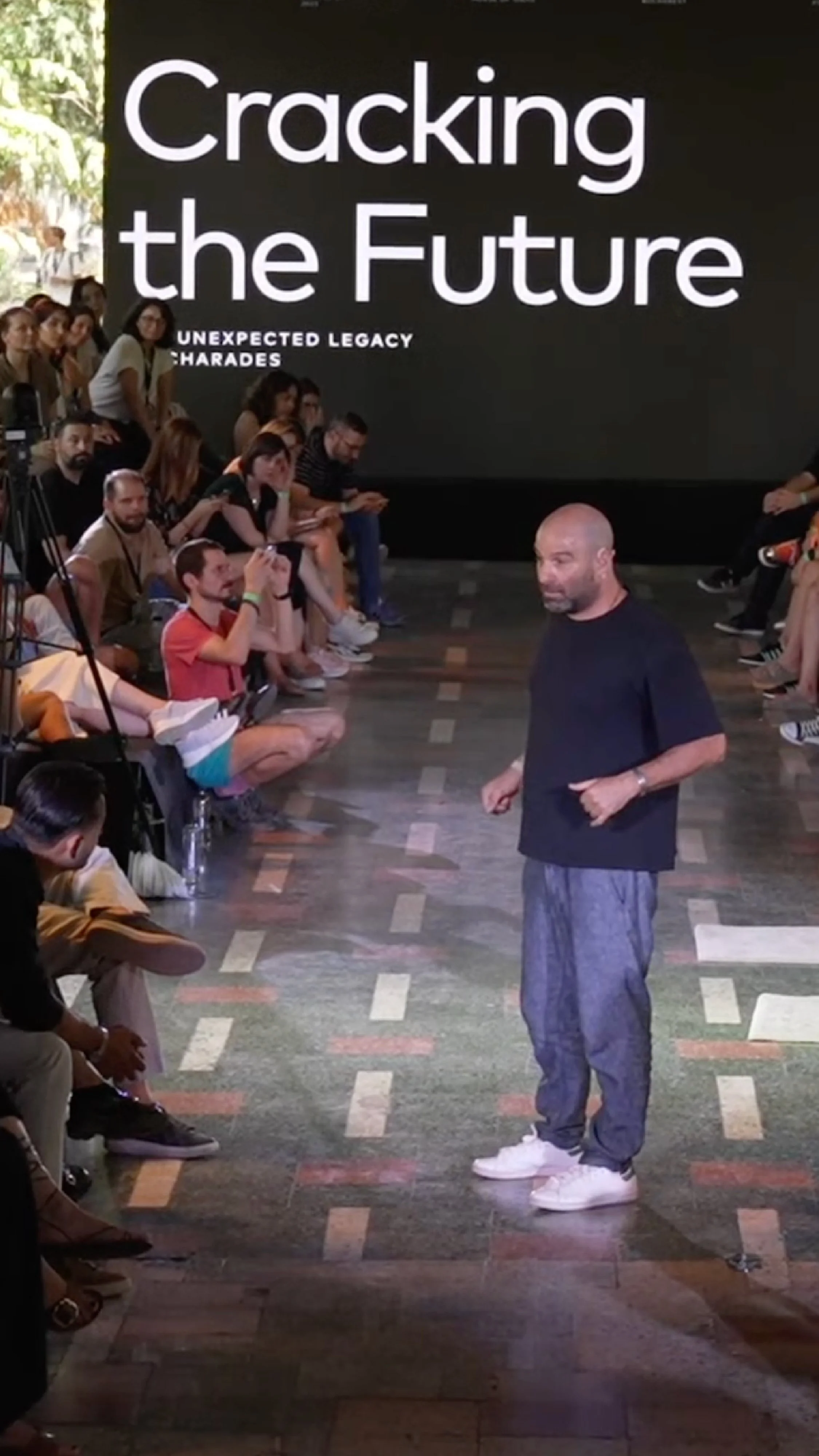 Topaz Adizes standing in front of an audience during a presentation with a large screen behind him that reads 'Cracking the Future.' The audience is seated on both sides, attentively listening and some taking photos.