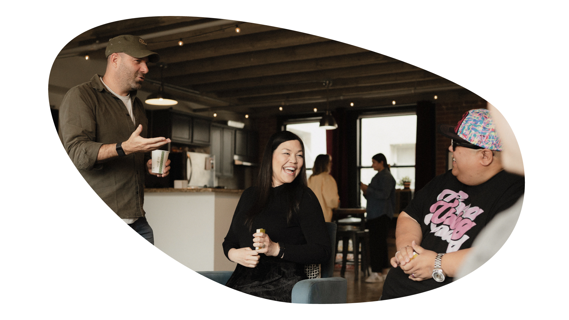 Three people in a cozy, modern indoor space are engaged in a lively conversation, smiling and laughing, with a kitchen in the background.