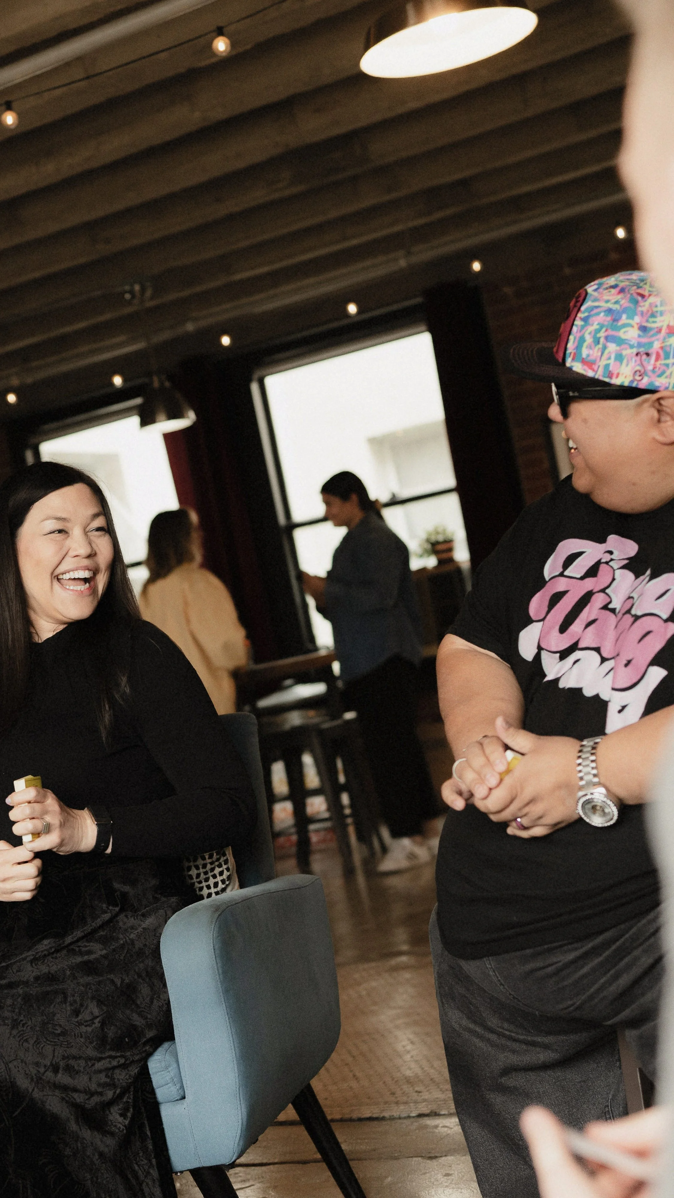 People enjoying a social gathering indoors, smiling and talking in a cozy, warmly lit room with wooden ceiling and large windows.