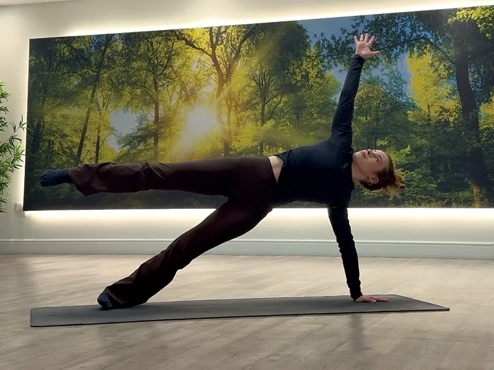 A woman practicing pilates indoors, performing a side plank pose with her right hand and left foot supporting her body, left arm raised overhead, against a backdrop of a forest scene with sunlight filtering through trees.