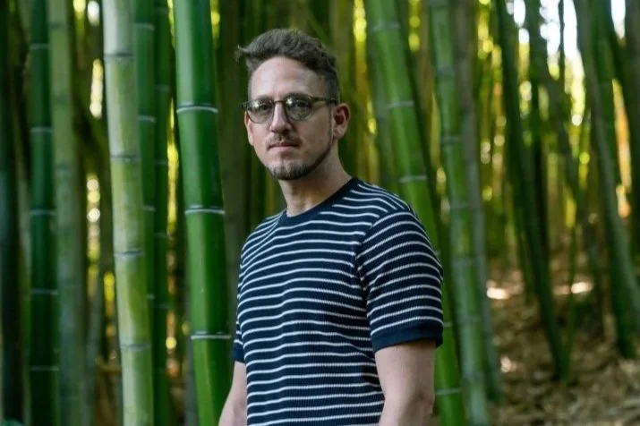 A man with glasses and a striped shirt stands in a bamboo forest.