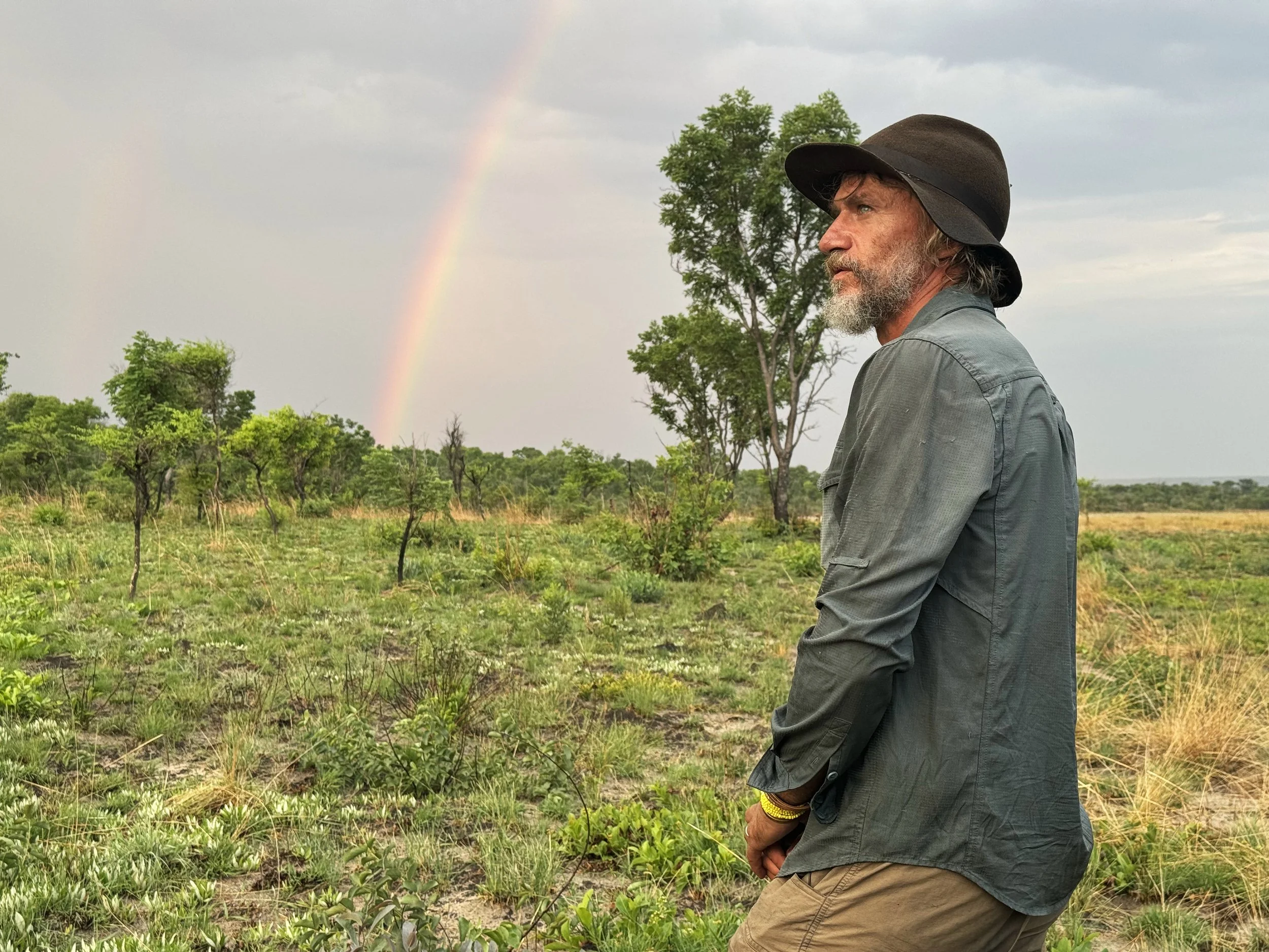 A man with a beard wearing a gray shirt and a wide-brimmed hat gazes at a rainbow in a grassy, sparse landscape with green trees and a cloudy sky.