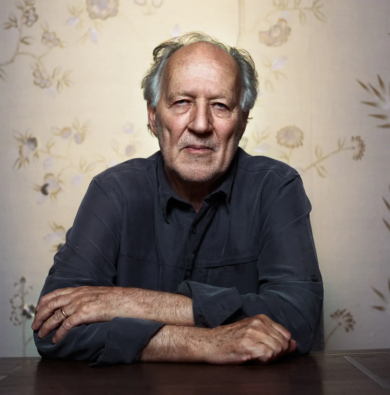 An older man with gray hair and a serious expression sitting at a table with folded arms, wearing a dark shirt, against a floral-patterned wallpaper background.