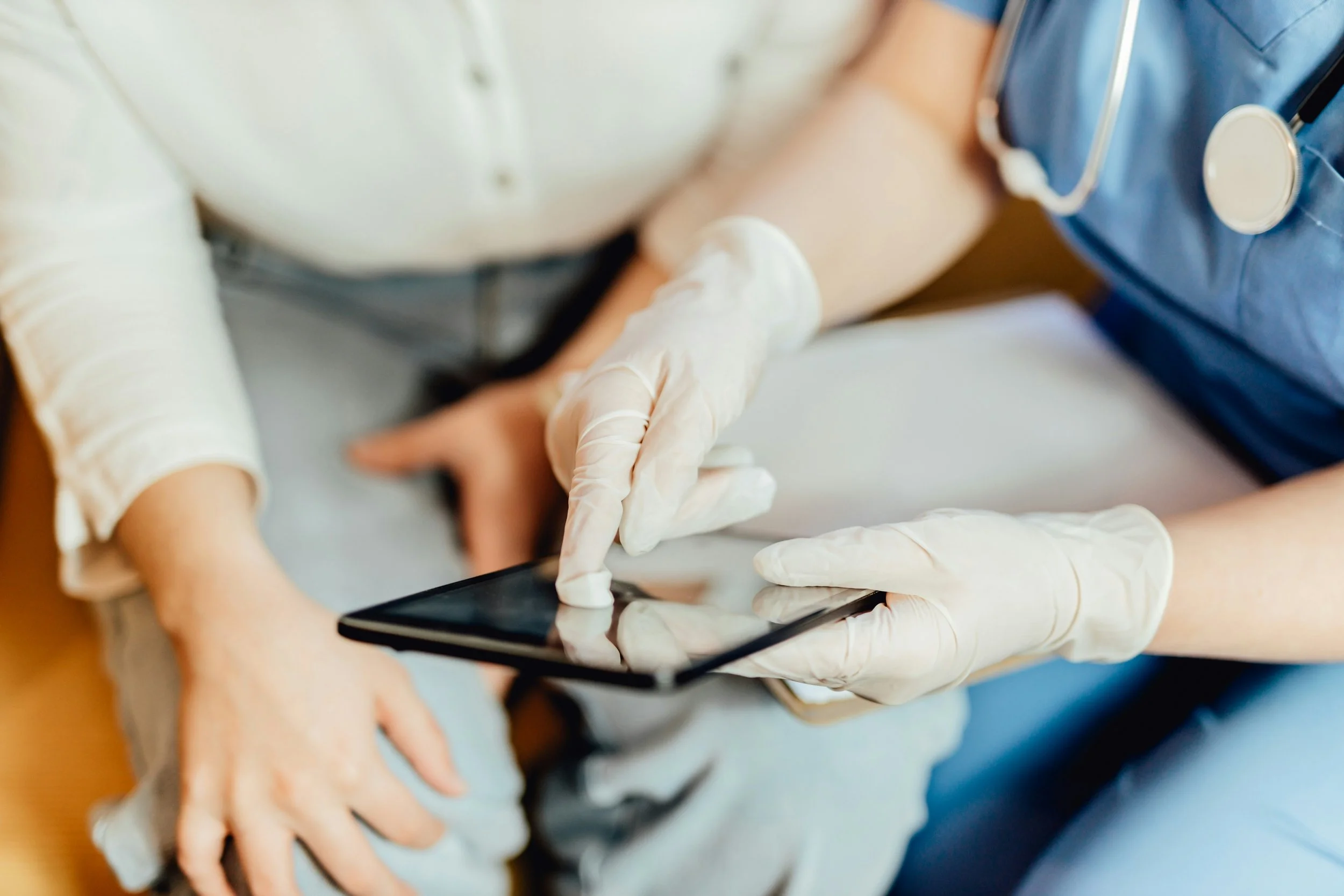 A healthcare worker in gloves using a tablet while a patient arm is being examined or treated, with a stethoscope around the healthcare worker's neck.