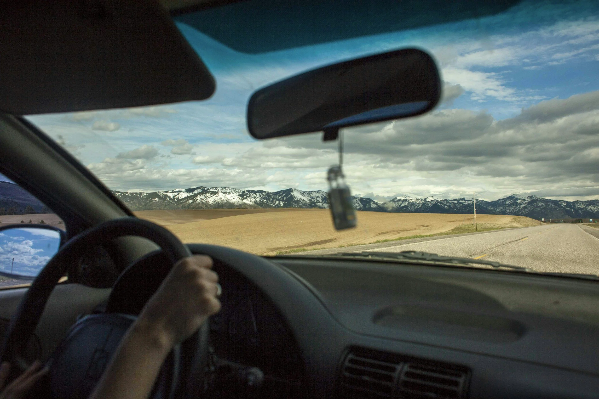 View from inside a car looking out through the windshield at a landscape with snow-capped mountains, cloudy sky, and open fields, while a person holds the steering wheel.