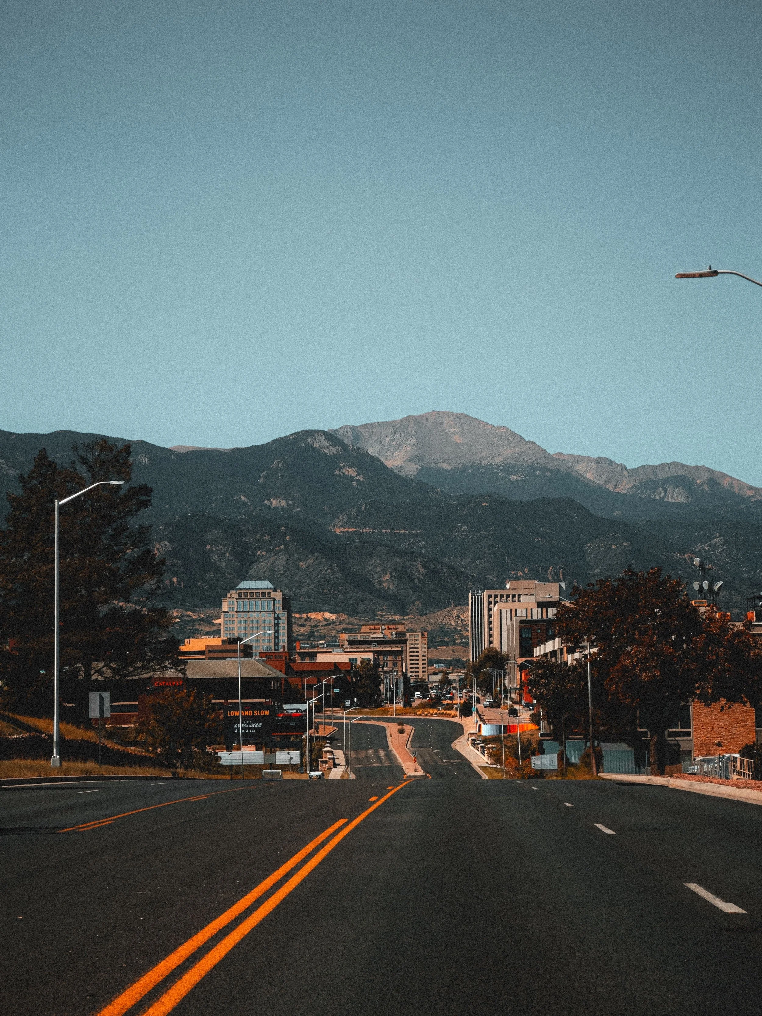 City street with buildings, mountains in background, and a clear blue sky, taken during dusk or dawn.