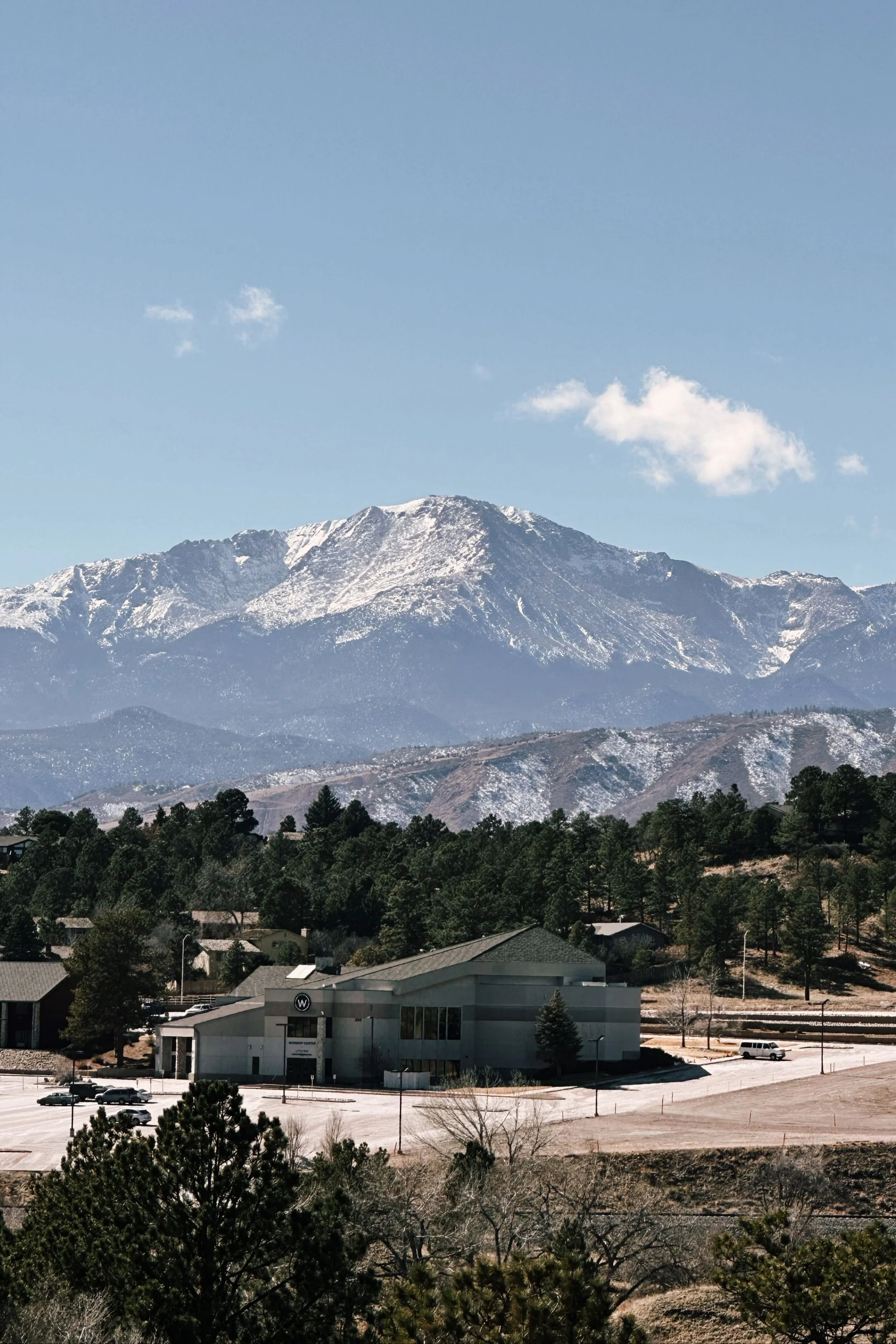 Snow-capped mountain range in the background with a small town and buildings in the foreground, under a clear blue sky with a few clouds.