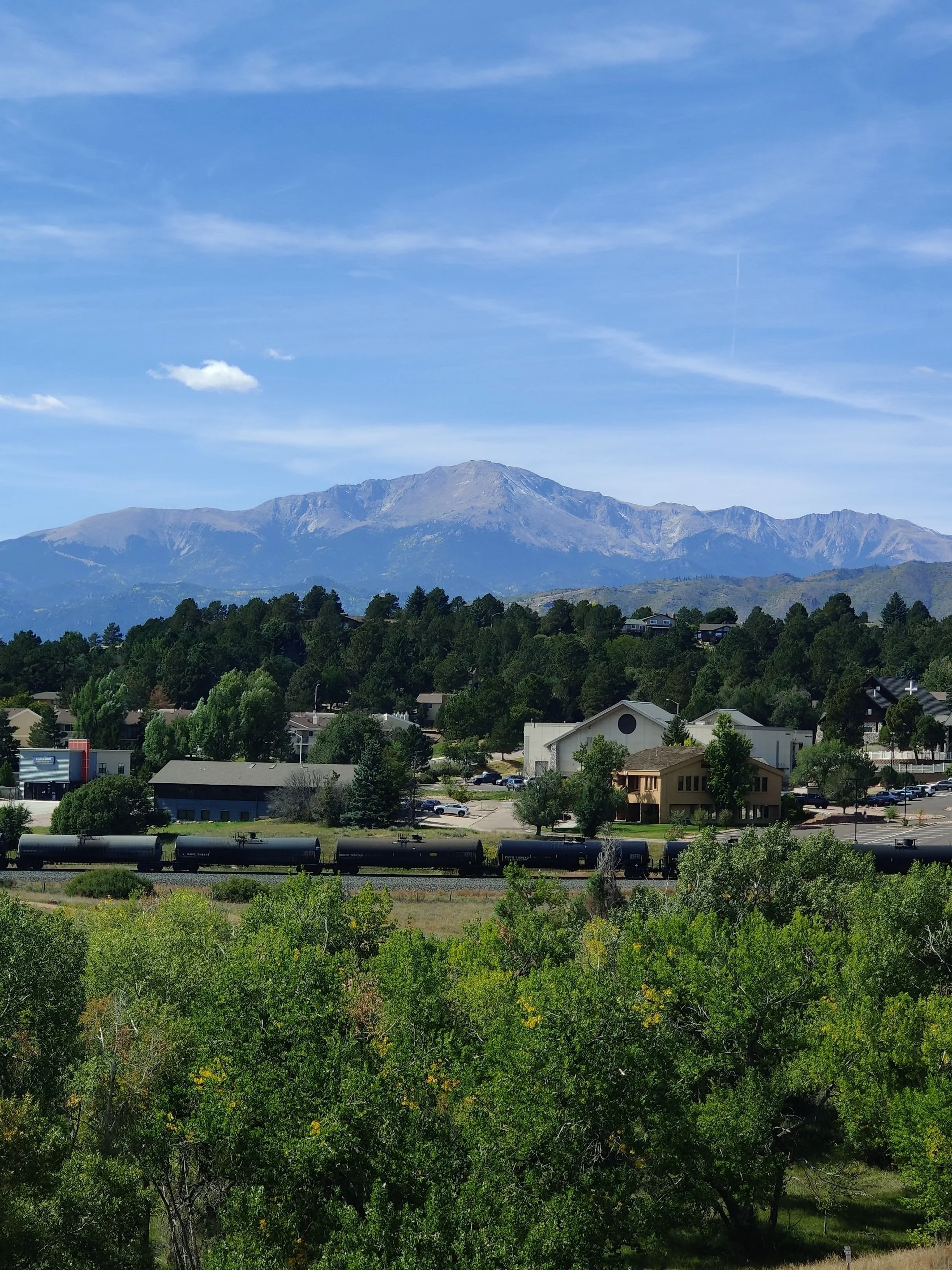 View of a mountainous area with houses and trees in the foreground, and a train on tracks in the lower part of the image under a partly cloudy sky.