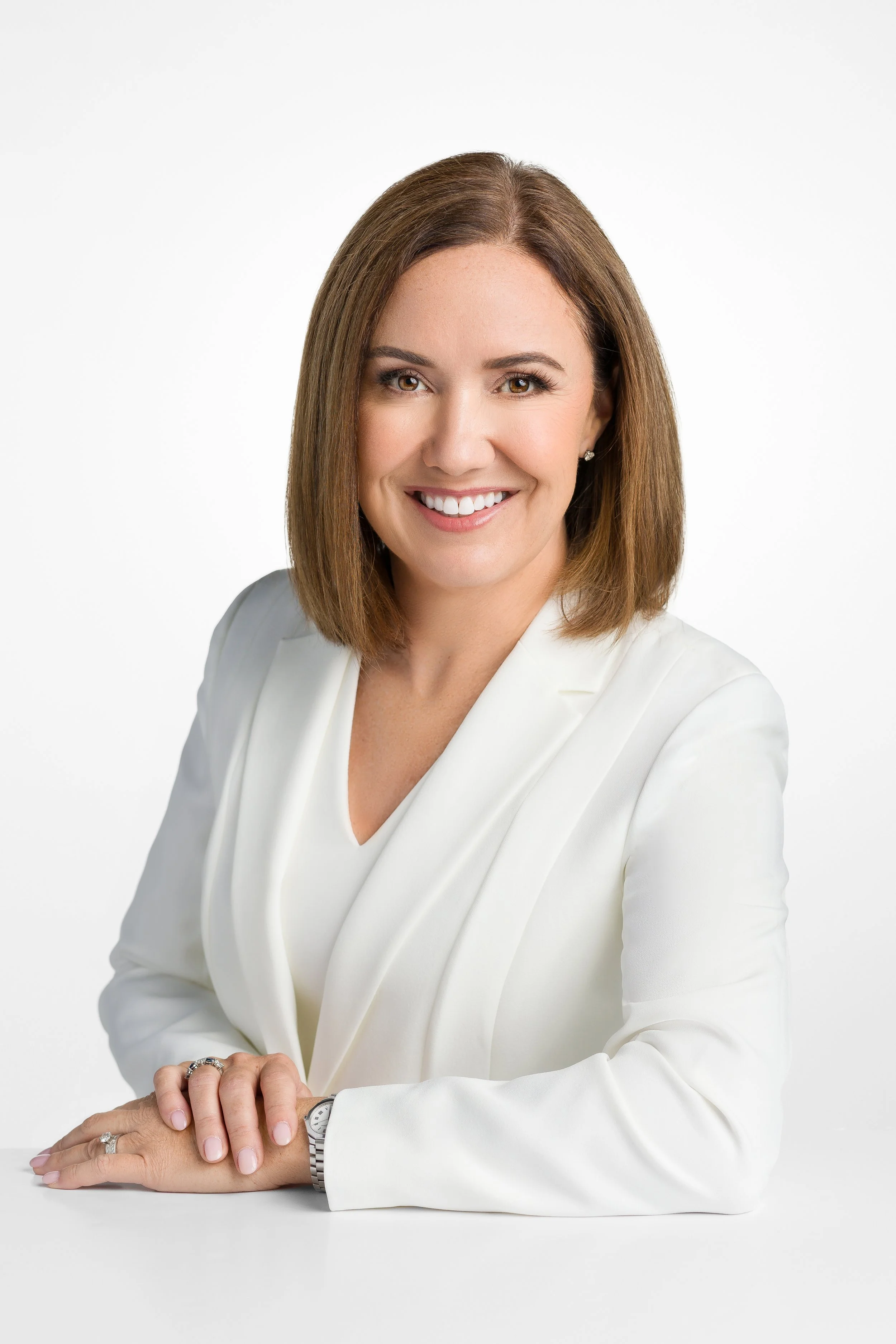 Portrait of a woman with shoulder-length brown hair wearing a white blazer, smiling, posed with her hands resting on a white surface with a white background.