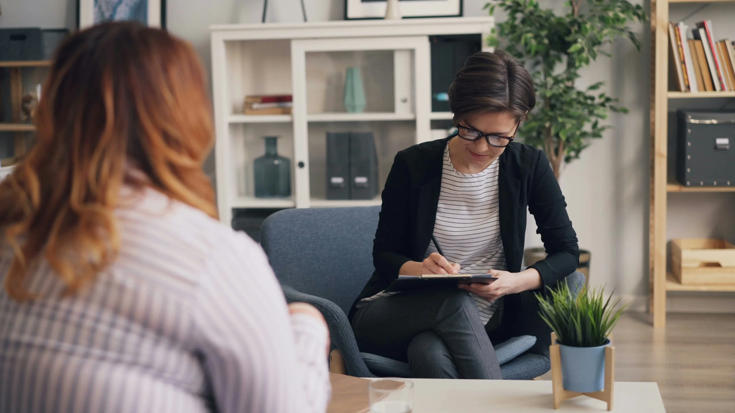 Two women sitting in a modern office space, one is taking notes on a clipboard while the other, wearing glasses and a striped shirt, is listening attentively. Shelves with books and decorative items are in the background.