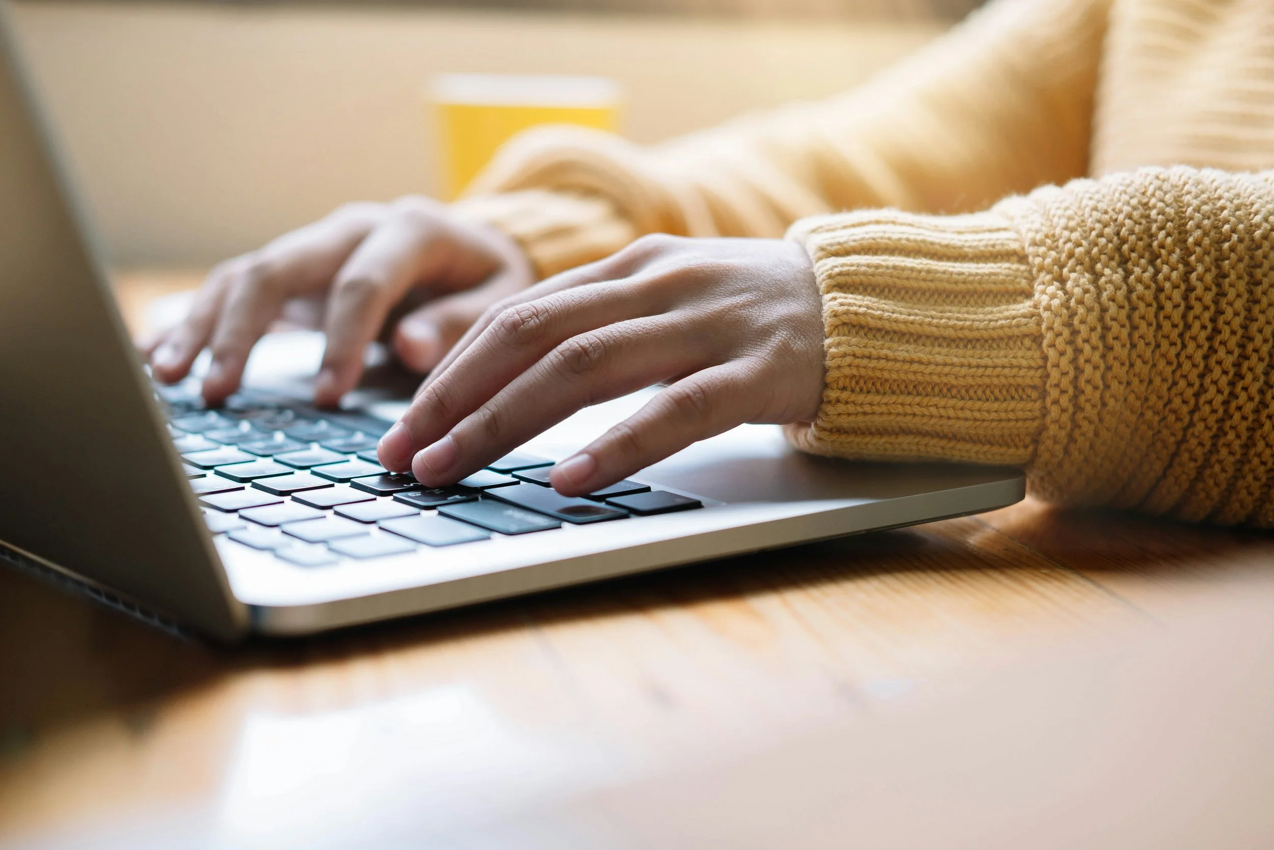 Person wearing a yellow sweater typing on a laptop keyboard with a yellow mug blurred in the background.