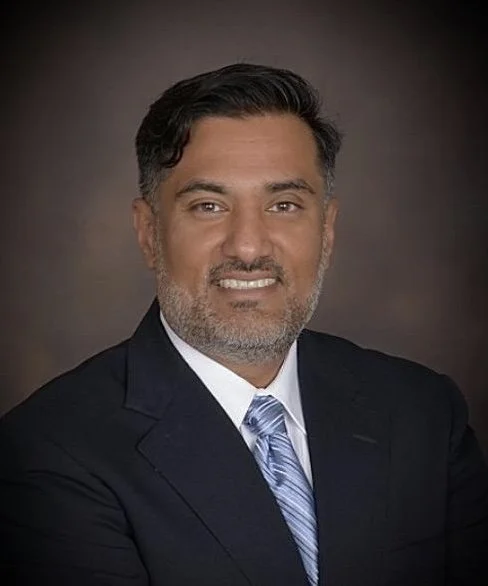 Professional headshot of a man in a dark suit, light blue tie, with short dark hair and beard, smiling against a dark background.