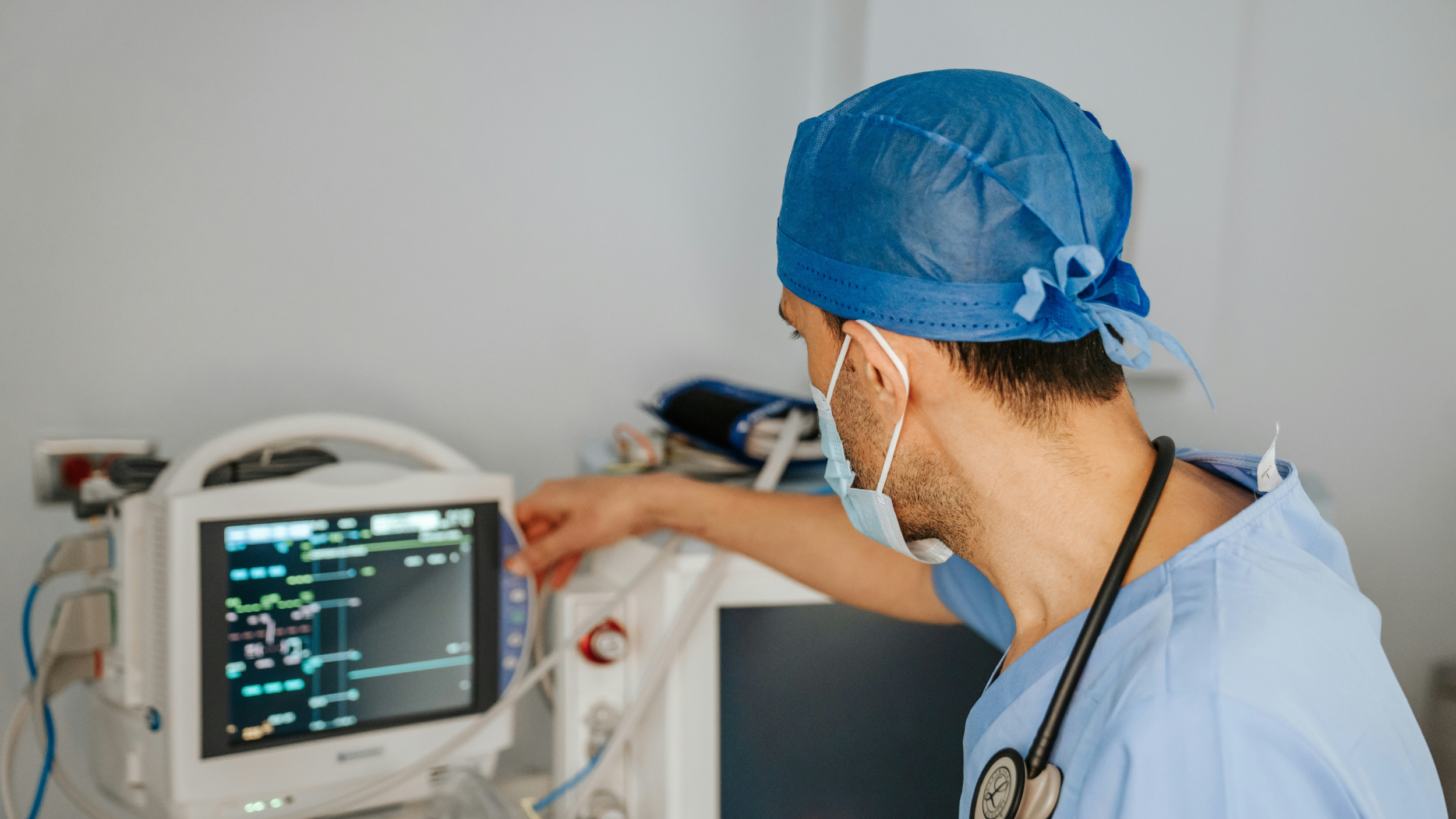 A clinician in scrubs monitors a patient's vital signs on a bedside monitor in a clinical setting.