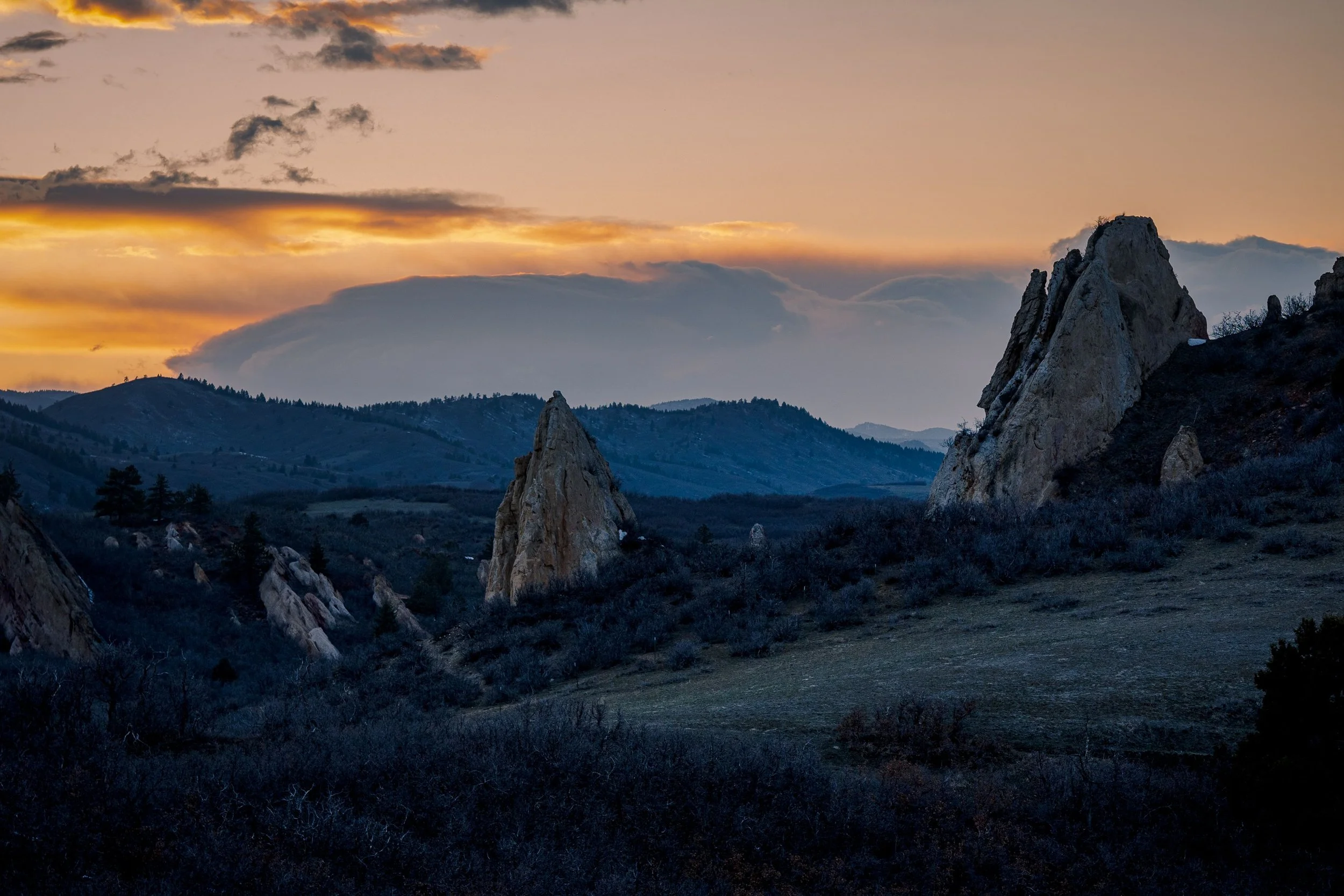 A scenic view of a mountain range during sunset with large rock formations in the foreground and layers of mountains extending to the horizon under a colorful sky.