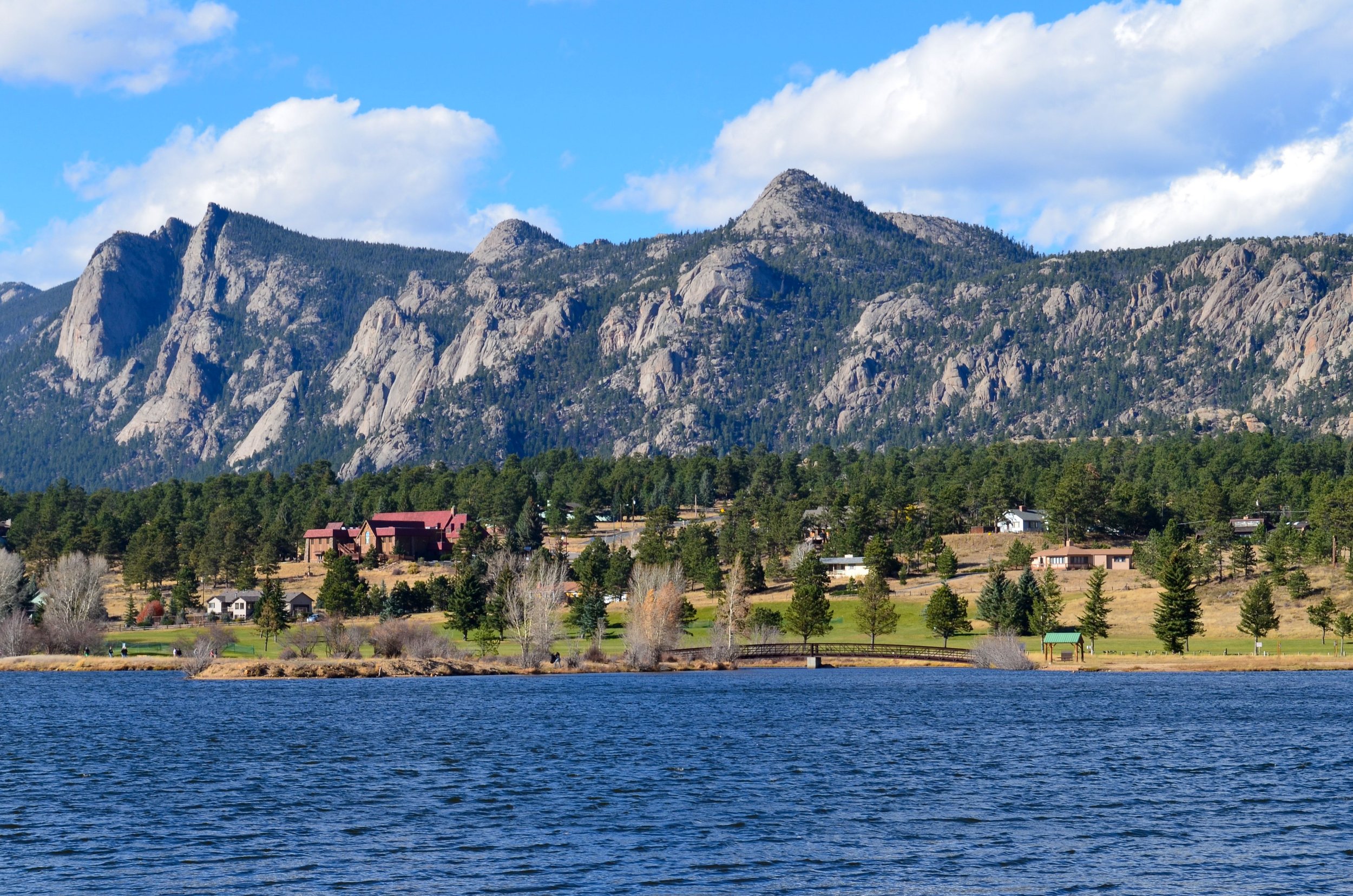 Scenic view of a mountain range with rocky peaks and dense forest, a small residential area with houses at the base, and a body of water in the foreground under a partly cloudy sky.