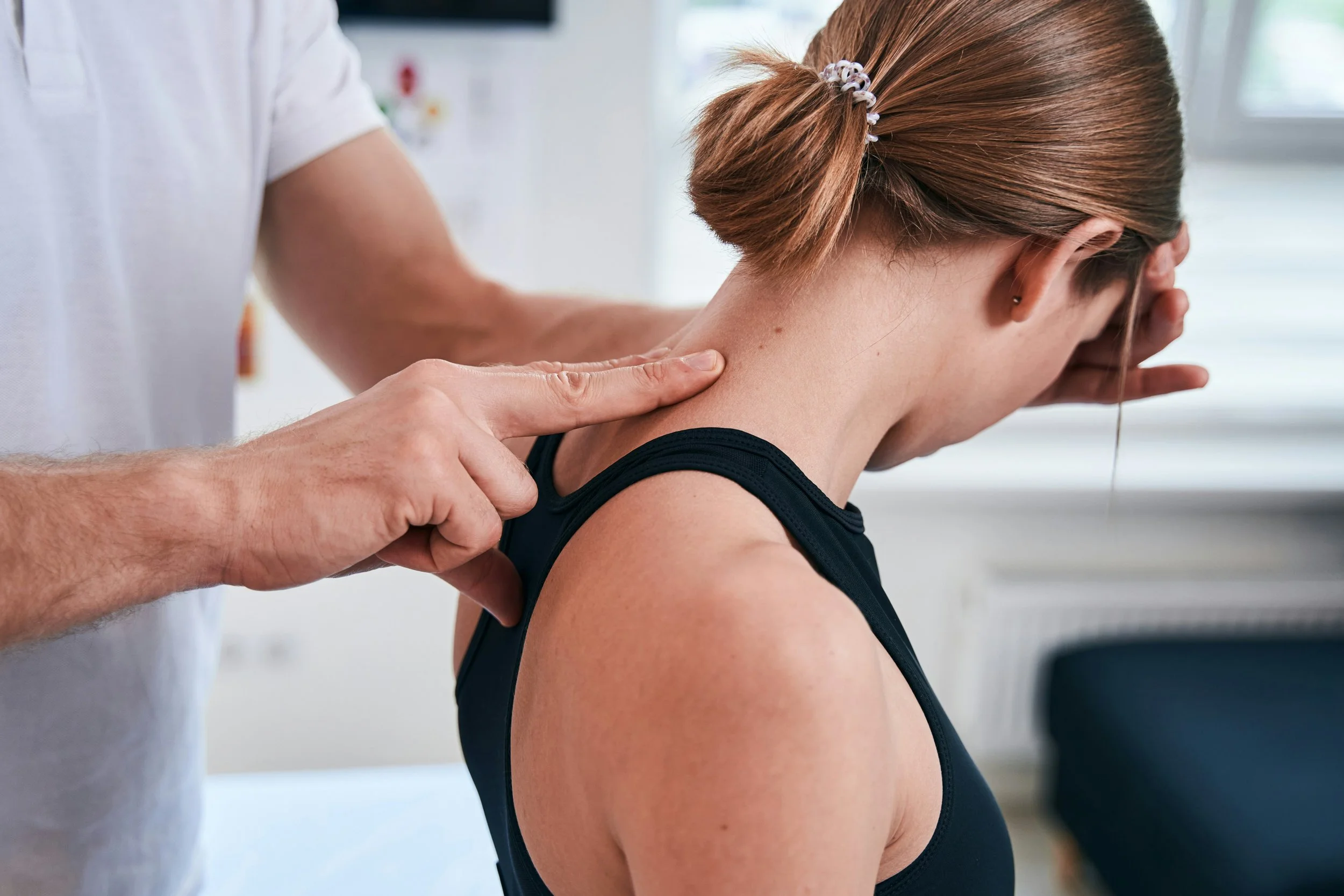 A person with red hair in a ponytail, wearing a black tank top, is receiving a chiropractic adjustment or neck therapy from a healthcare professional in a clinical setting.